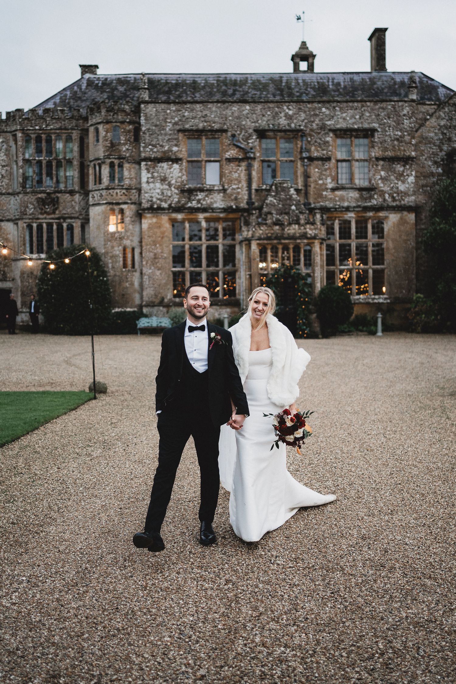 A couple in formal wedding attire poses on a gravel driveway in front of a grand historic stone manor house.