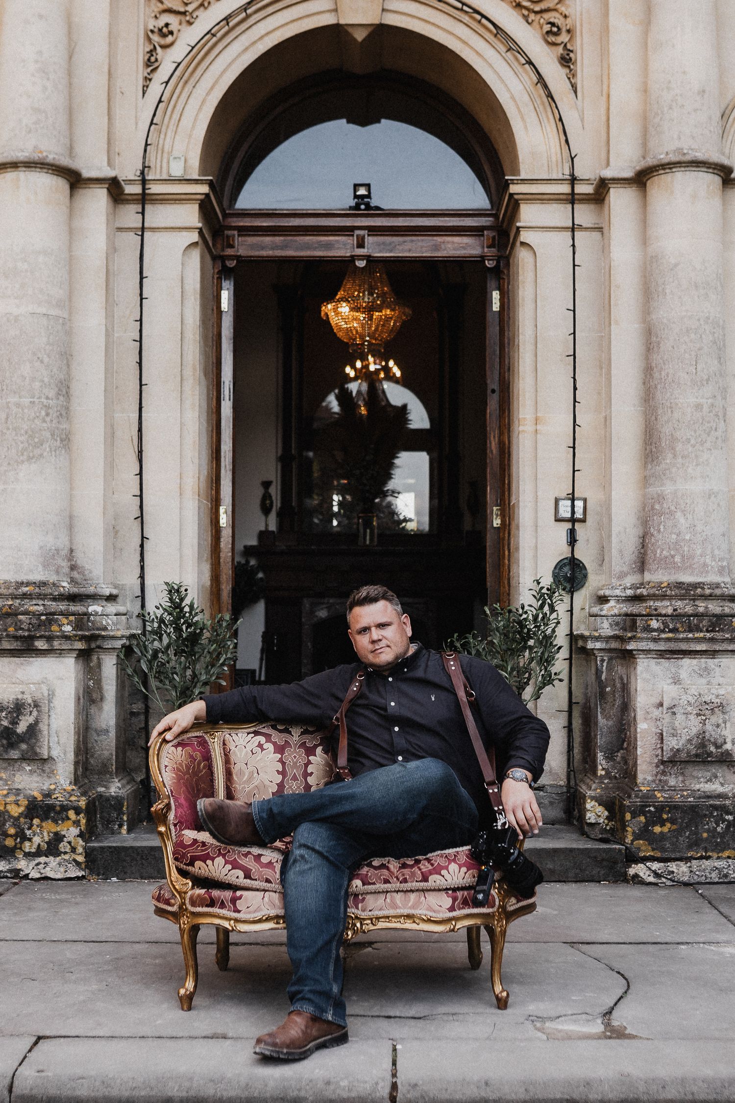 An antique floral sofa sits in front of an ornate stone archway entrance with a golden chandelier visible inside.
