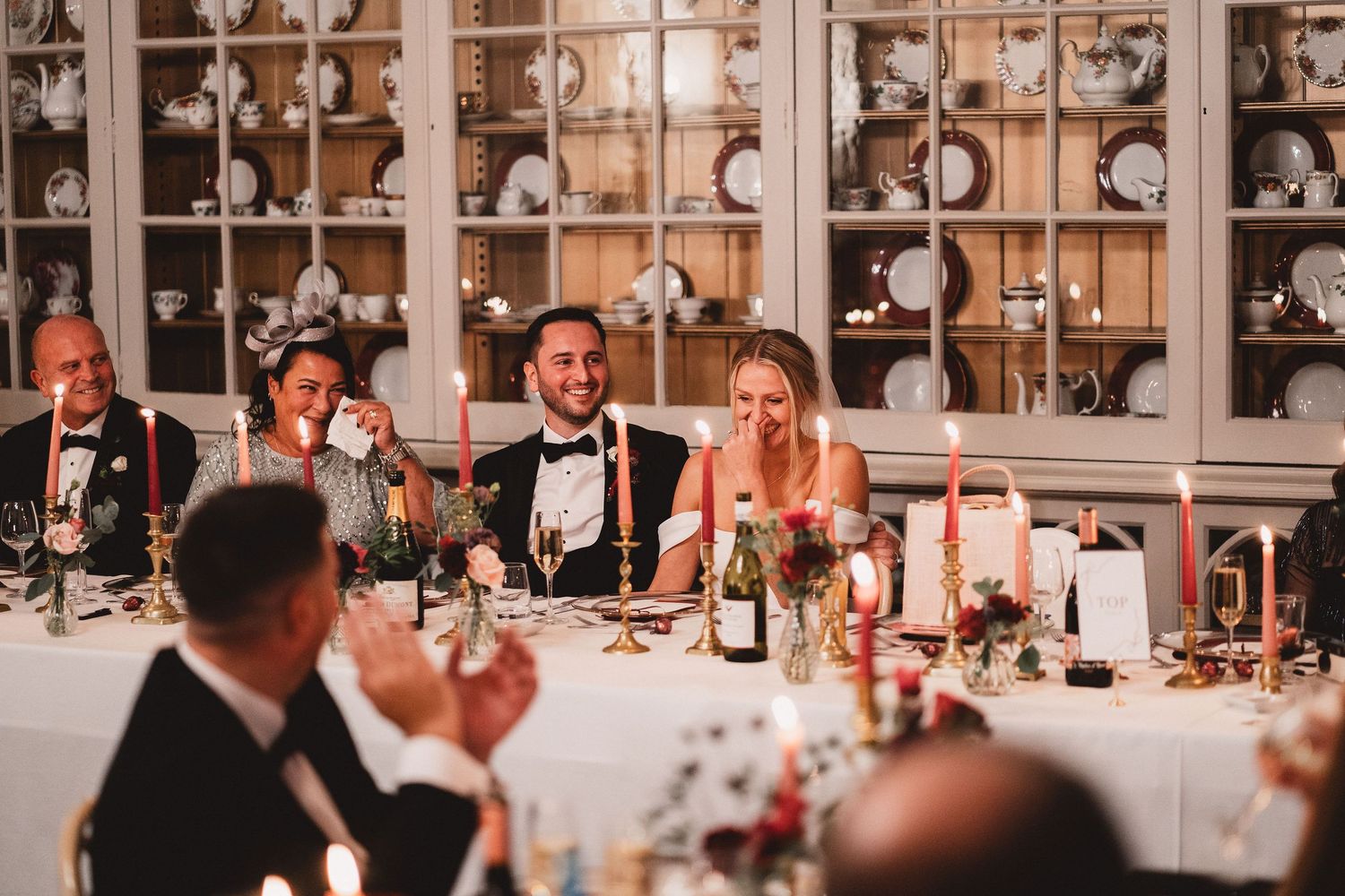 Wedding guests seated at an elegant dinner table with candles and wine glasses in front of illuminated display cabinets.