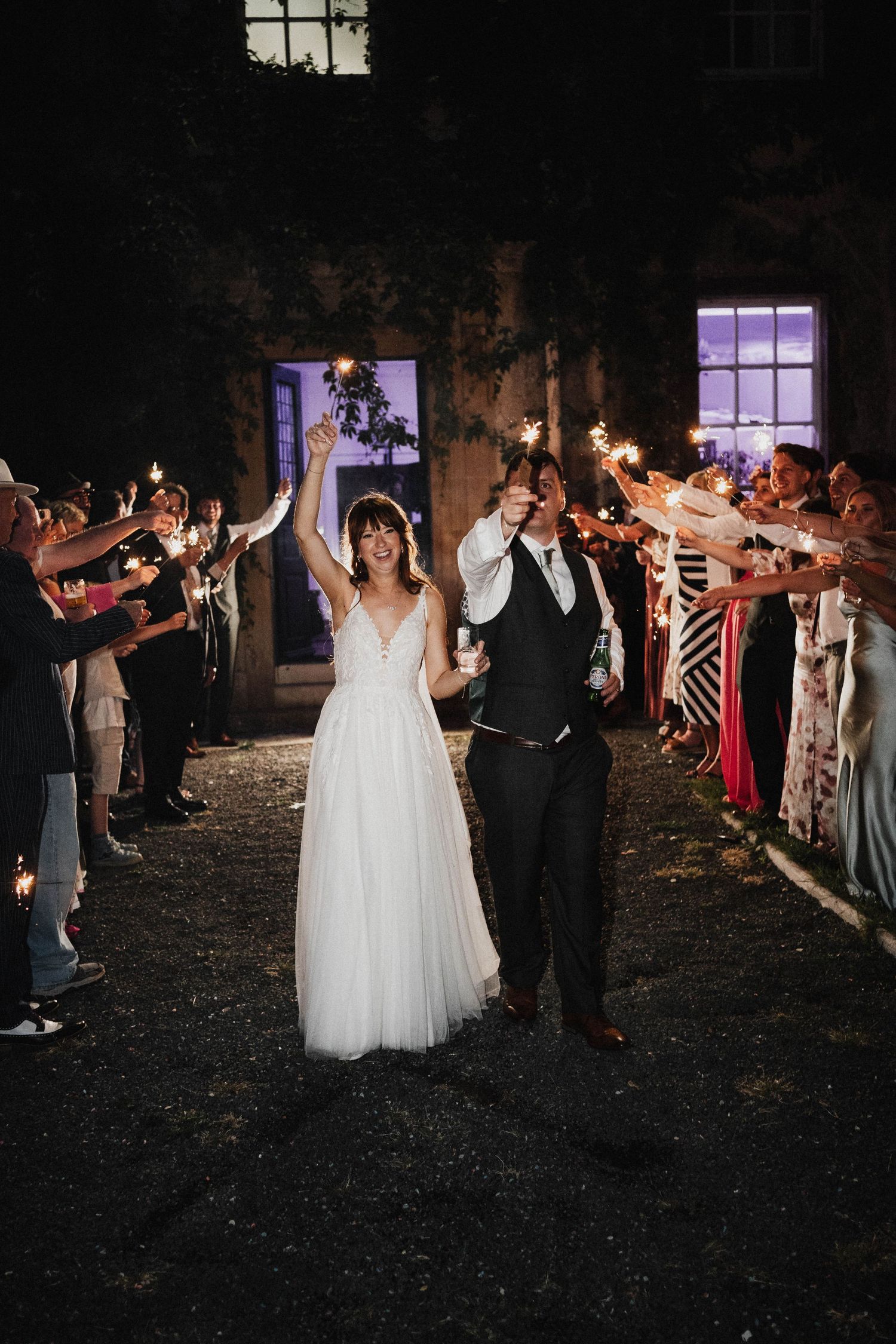 Wedding couple exits their reception at night through a sparkler tunnel lined with cheering guests.