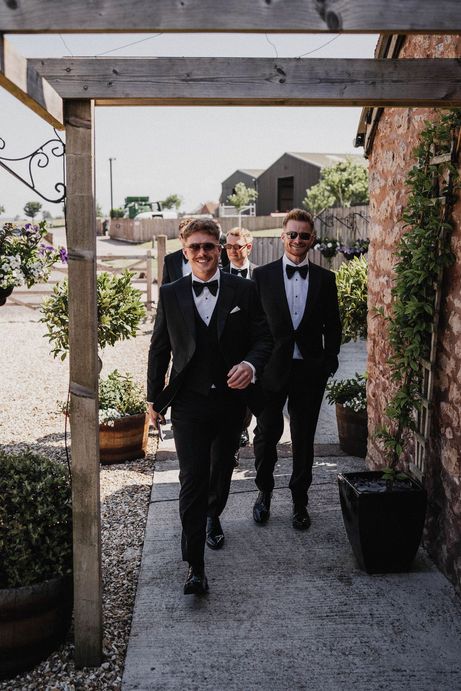 Two people in formal black tuxedos and bow ties walking under a wooden garden arbor decorated with potted plants.