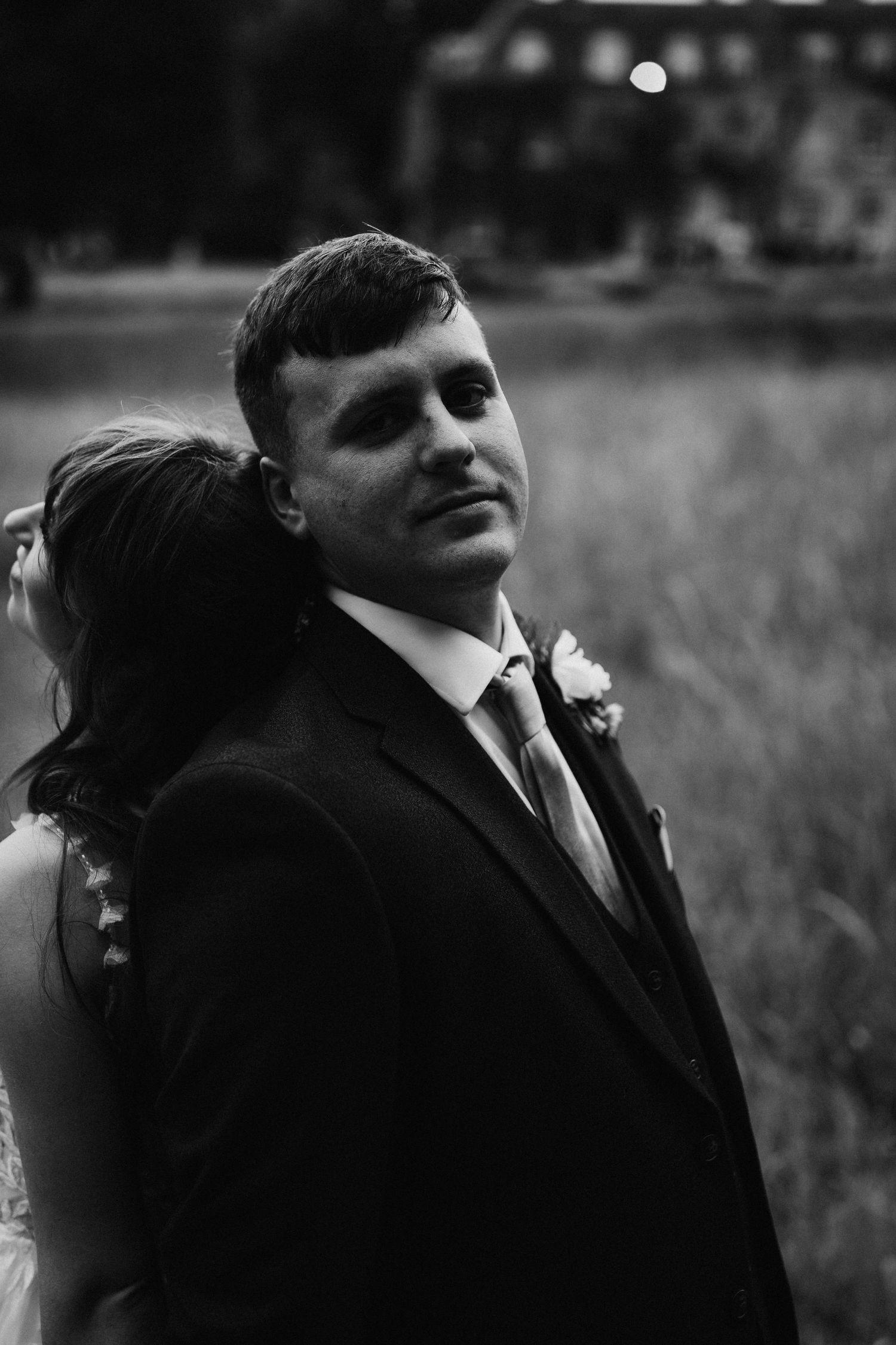 A black and white portrait of a person in a formal suit with a boutonniere at an outdoor wedding.