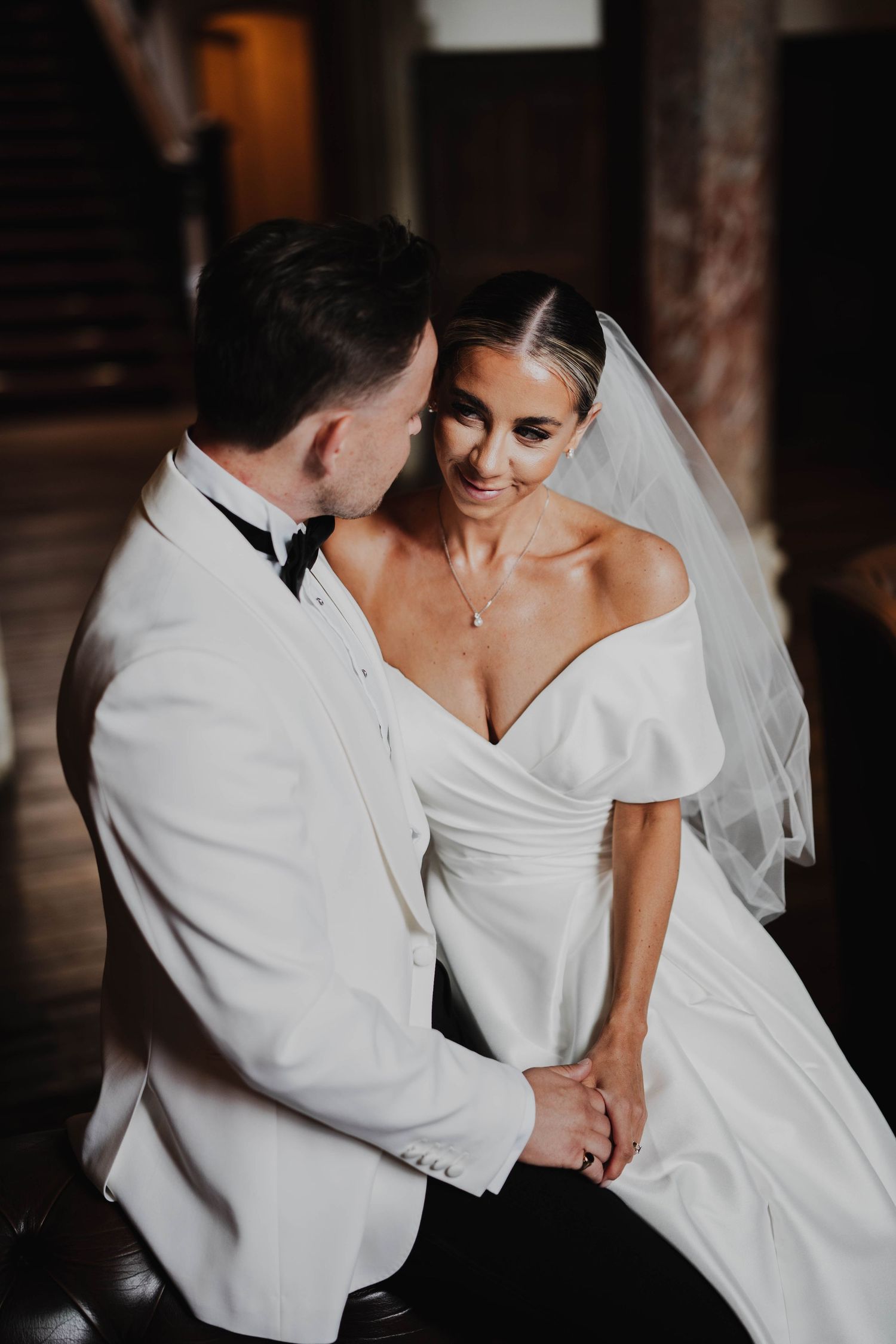 Bride in off-shoulder gown and veil smiles at groom in white tuxedo while holding hands indoors.