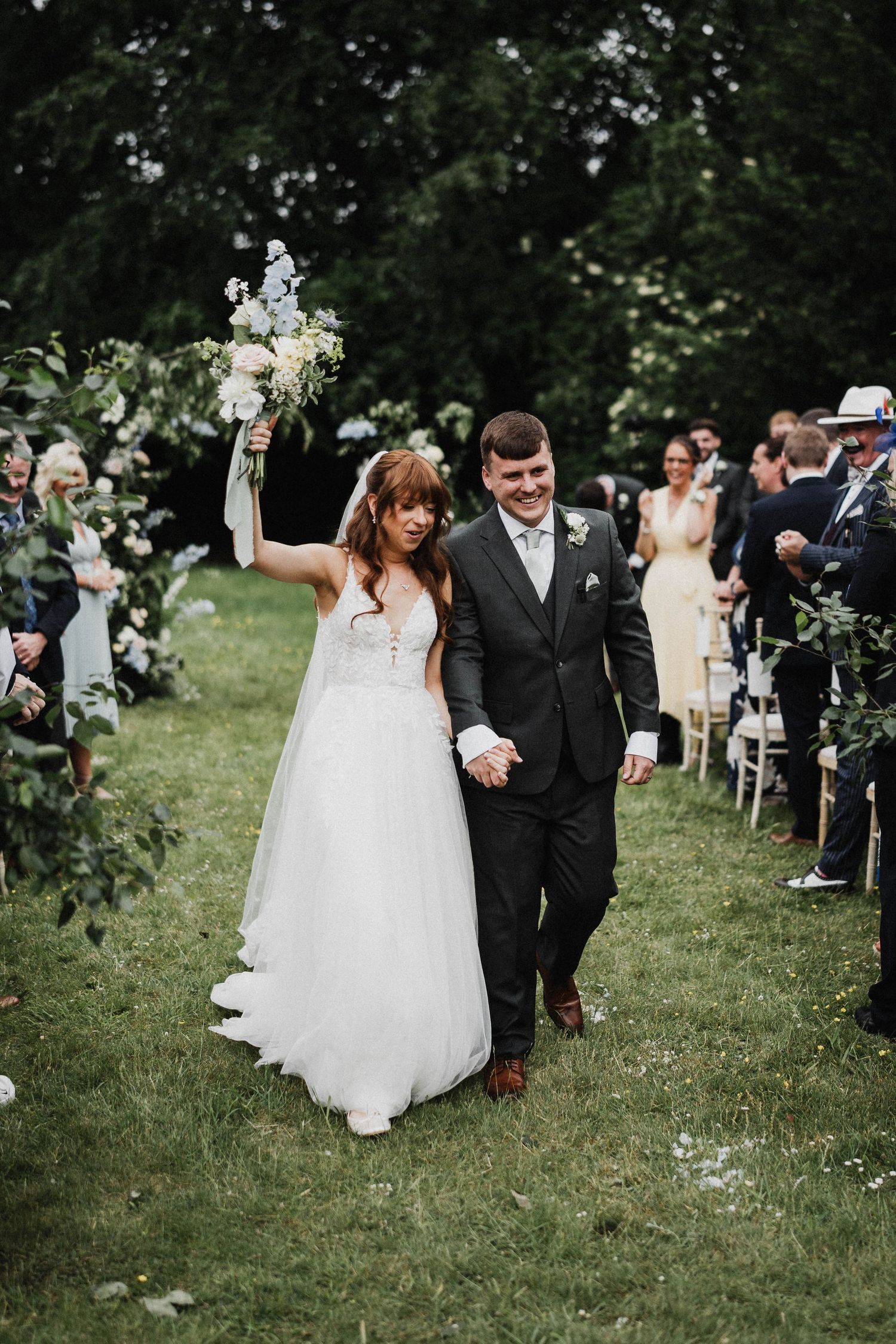 A couple walks down a tree-lined outdoor aisle as wedding guests look on during their garden ceremony.