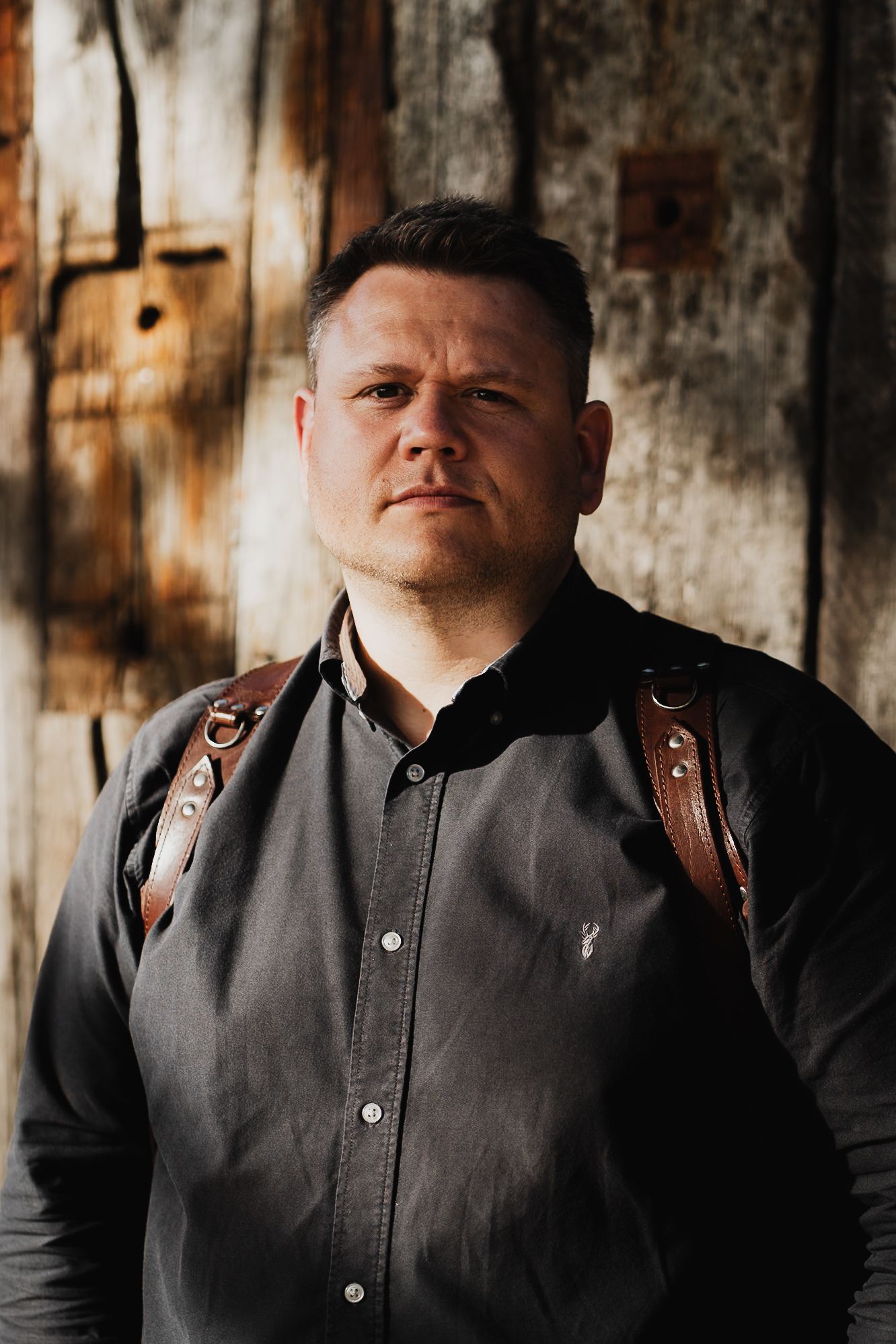 A serious man in a dark shirt with leather suspenders stands against a rustic wooden wall backdrop.