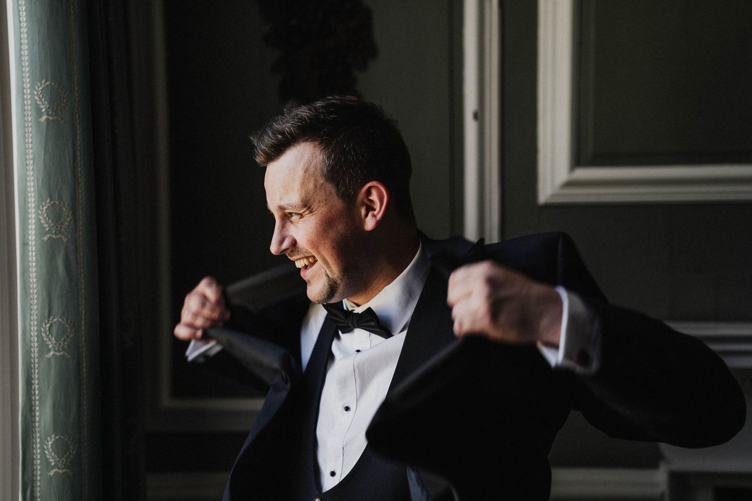 A person in a black tuxedo and bowtie clenches their fists and makes an energetic expression near a window.