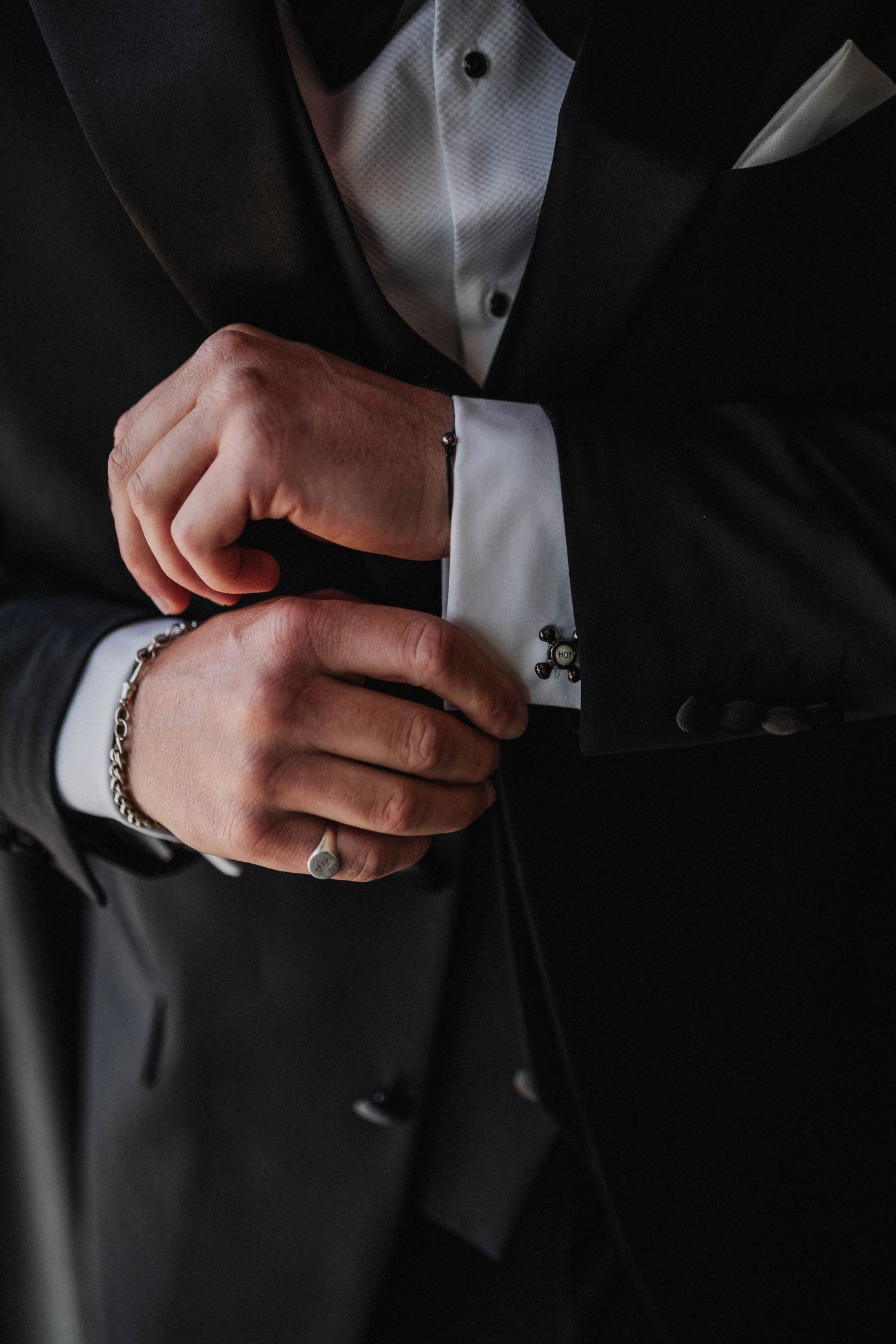 Close-up details of a black suit jacket with white dress shirt and silver chain accessory in moody lighting.