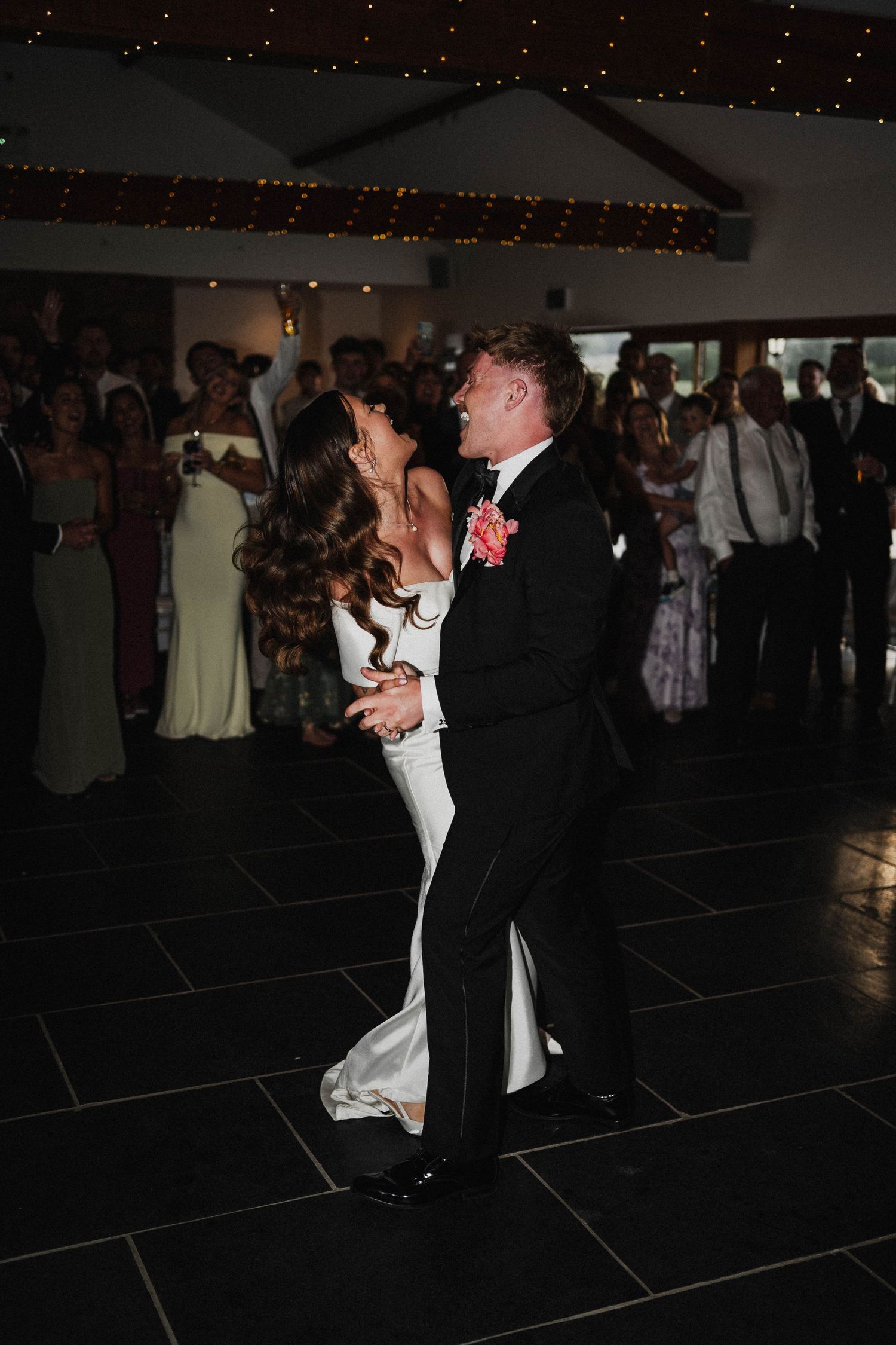 Bride and groom share their first dance on a dark tile floor under twinkling lights at their wedding reception.