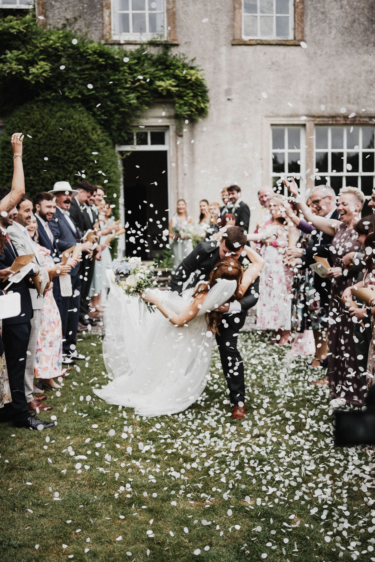 Wedding guests joyfully shower newlyweds with flower petals outside a historic stone house covered in climbing vines.