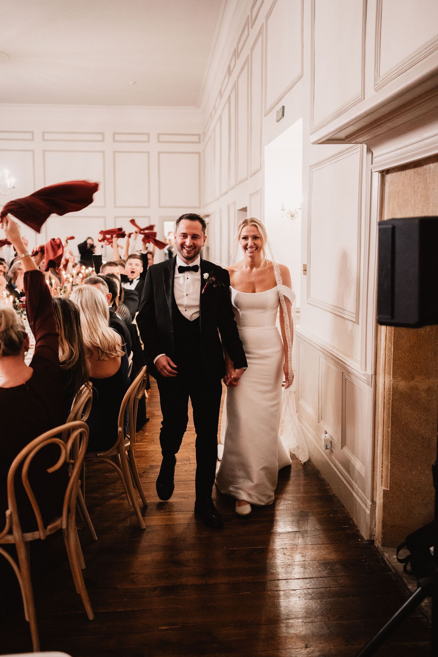 Wedding party walks down elegant hallway with white paneled walls and dark hardwood floors during reception celebration.