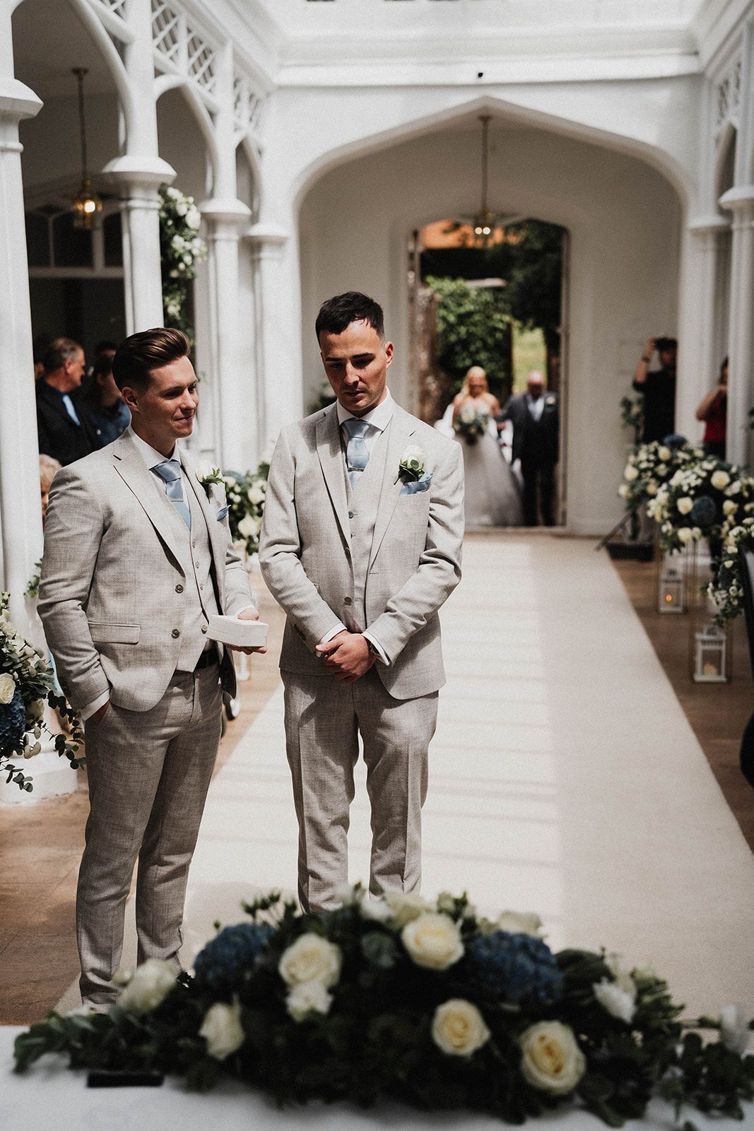 Groomsmen in light gray suits share candid moments in an elegant white columned hallway decorated with floral arrangements.
