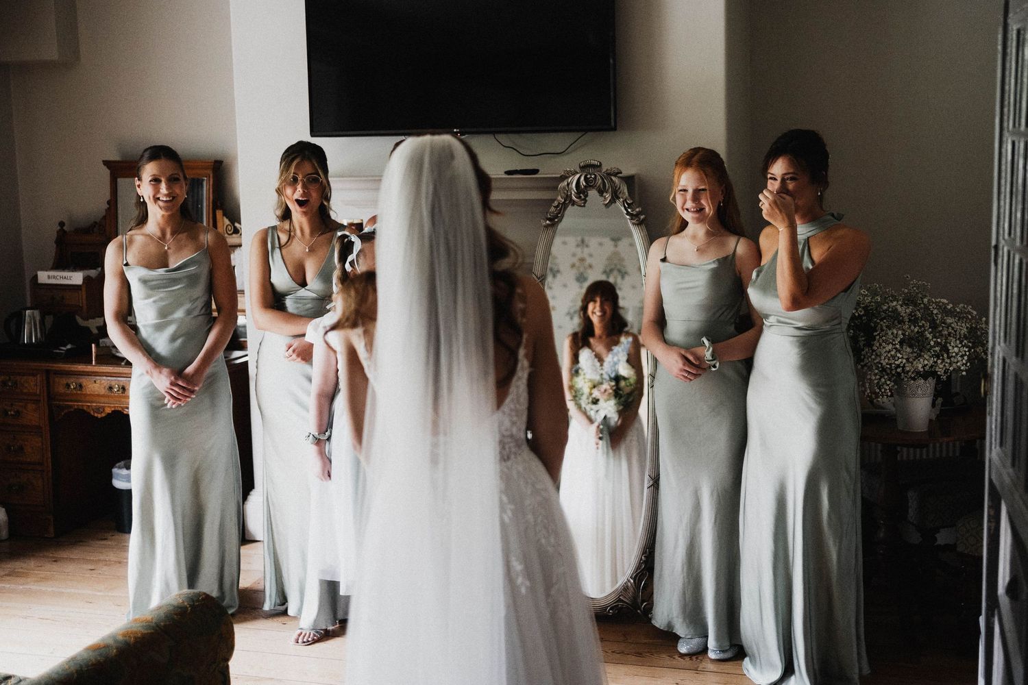 A group wearing light gray bridesmaid dresses stands together while one person adjusts a white bridal gown.