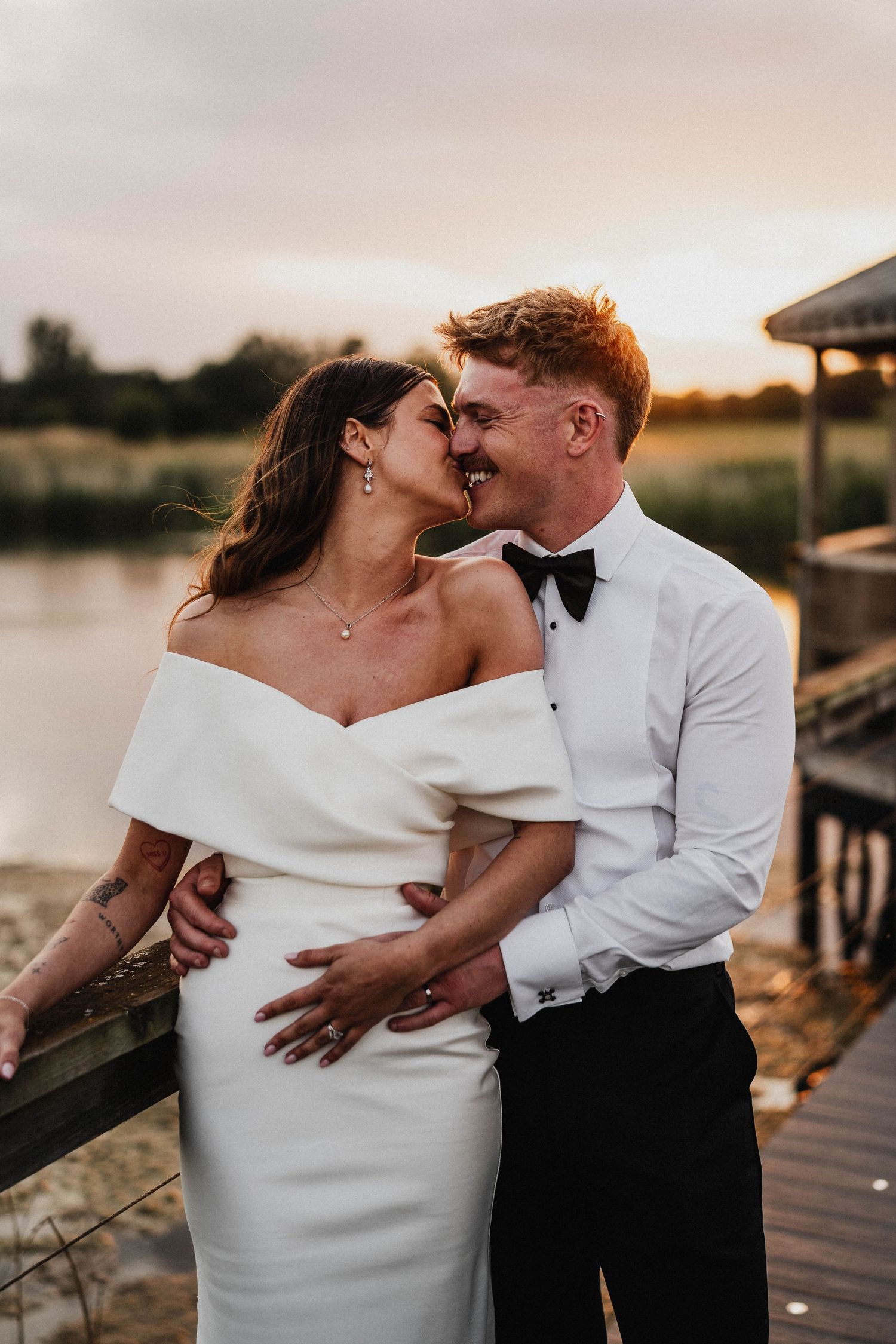 A romantic newlywed couple embraces on a wooden dock at sunset in their wedding attire.