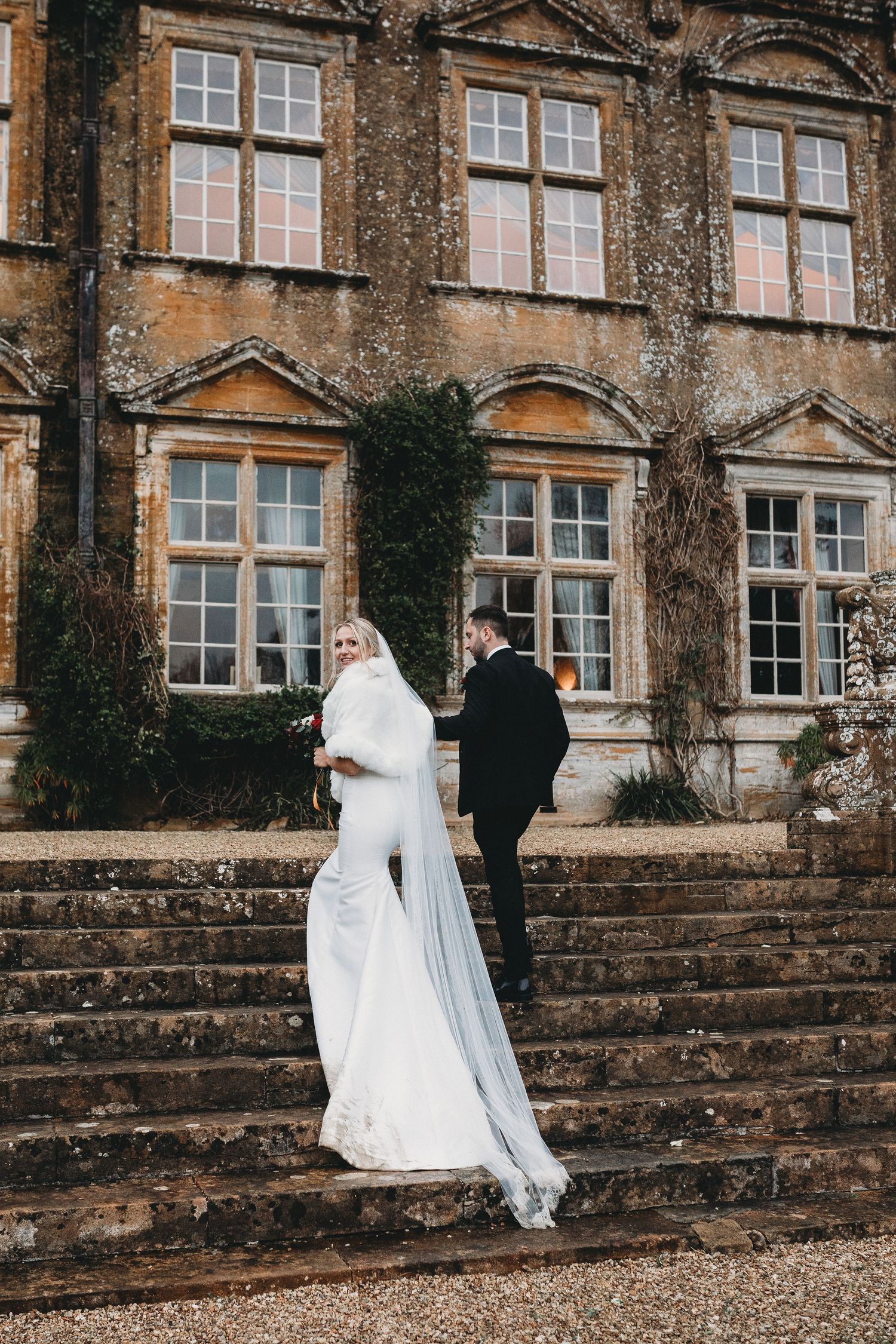 A couple in wedding attire walks along stone steps beside a grand historic building with ivy-covered walls.