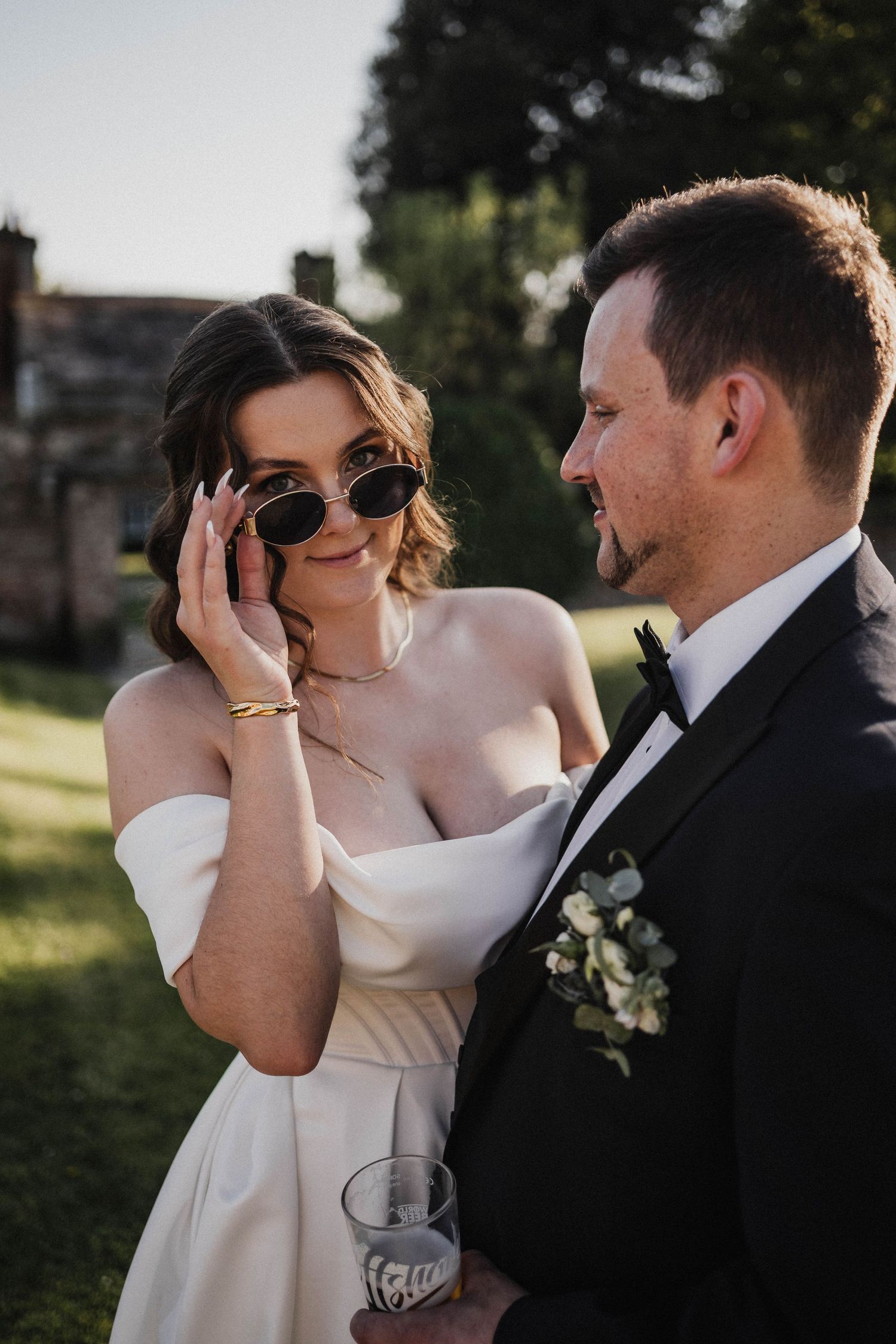 A bride in a white off-shoulder dress wearing sunglasses stands close to her groom in a black tuxedo during an outdoor wedding.