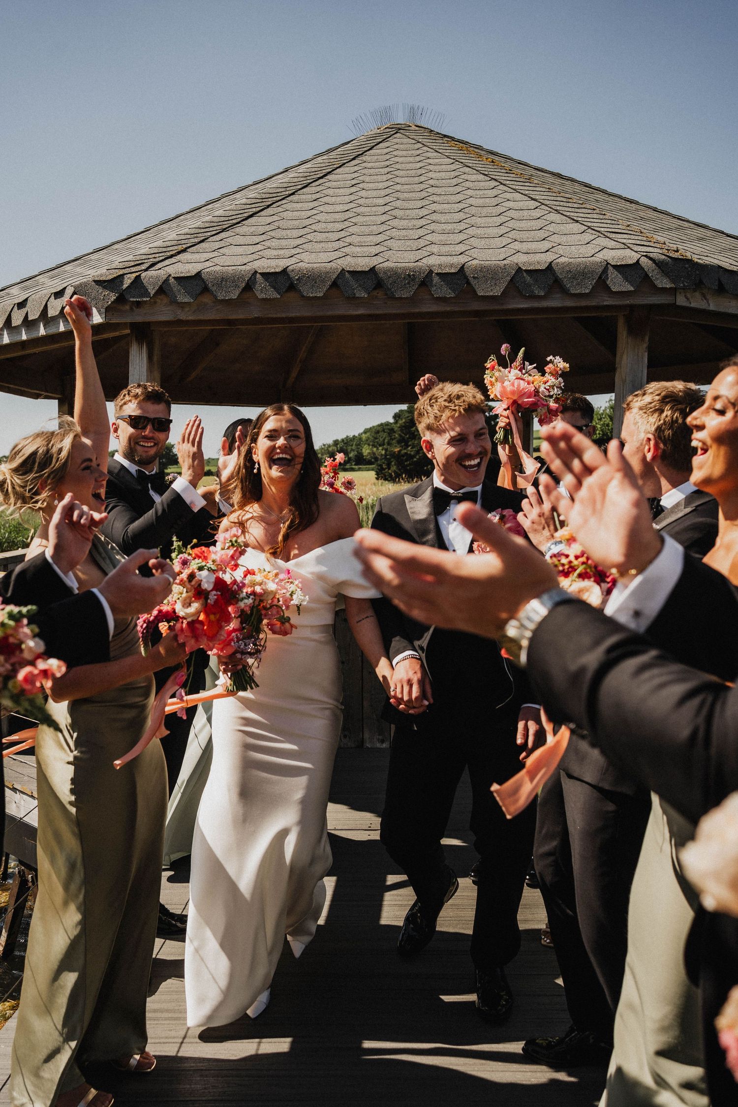Wedding guests form a lively dance line at an outdoor reception under a gazebo roof, creating a festive celebration moment.
