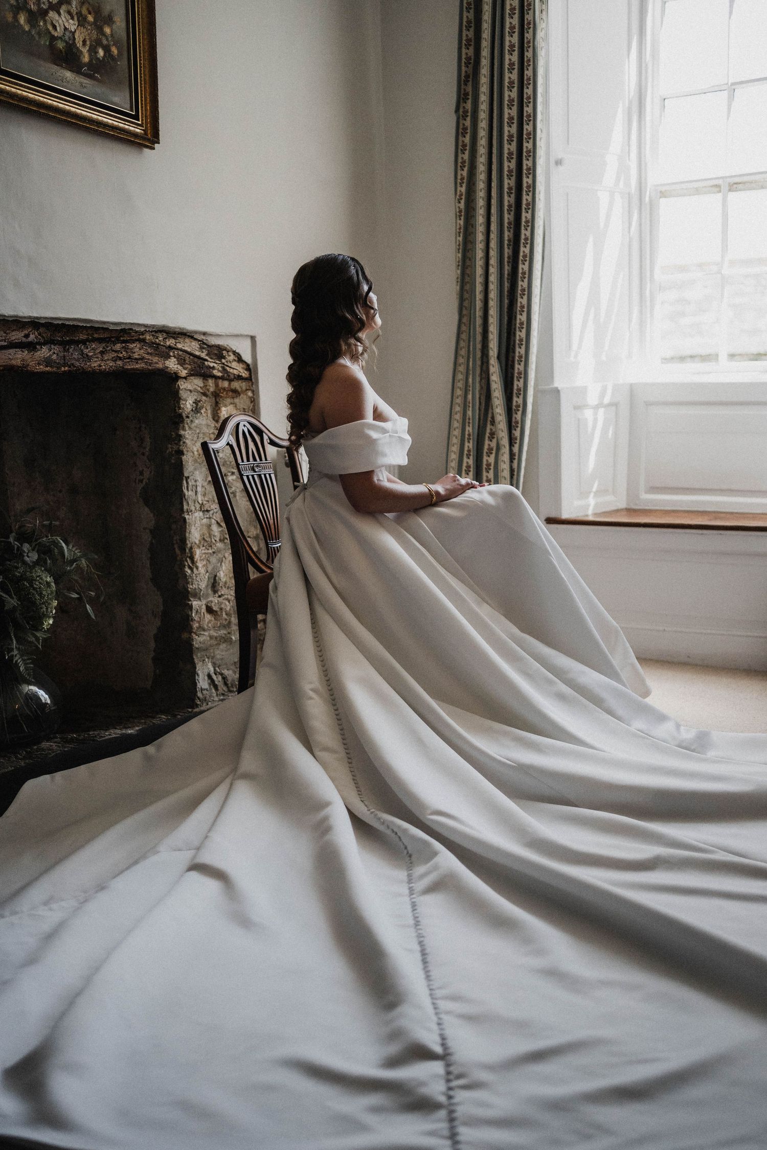 Elegant bride in white off-shoulder gown sits by window in vintage room with stone fireplace and flowing train.