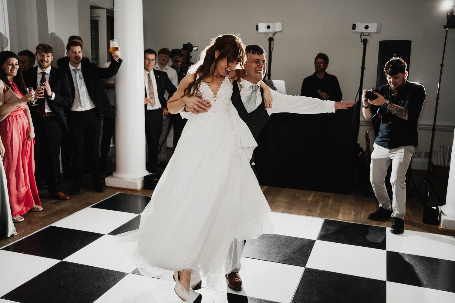 Two people dance together on a black and white checkered floor during a wedding reception while guests look on.