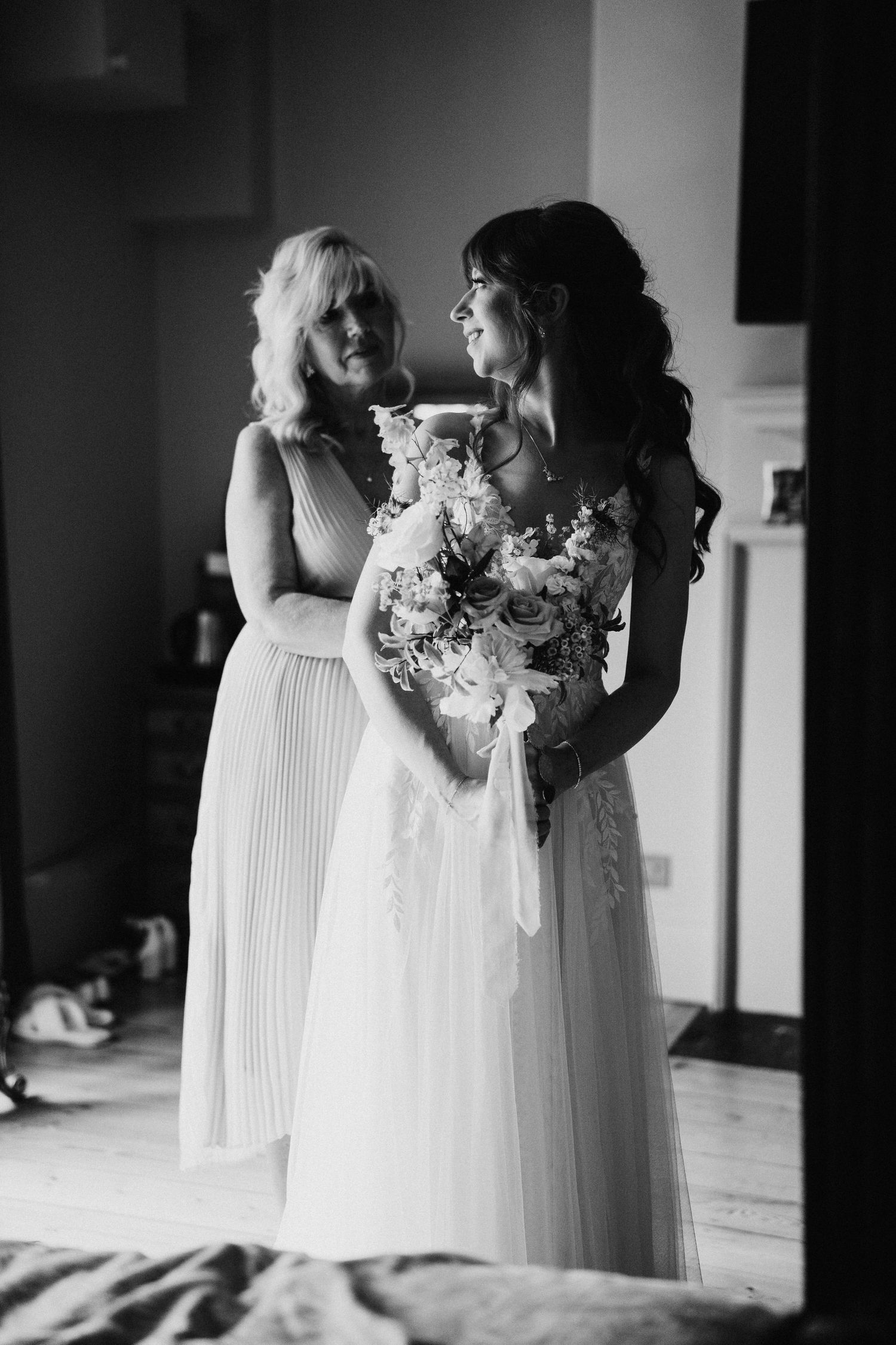 Black and white photo of a bride in a lace gown holding a floral bouquet while sharing a tender moment with bridesmaid.