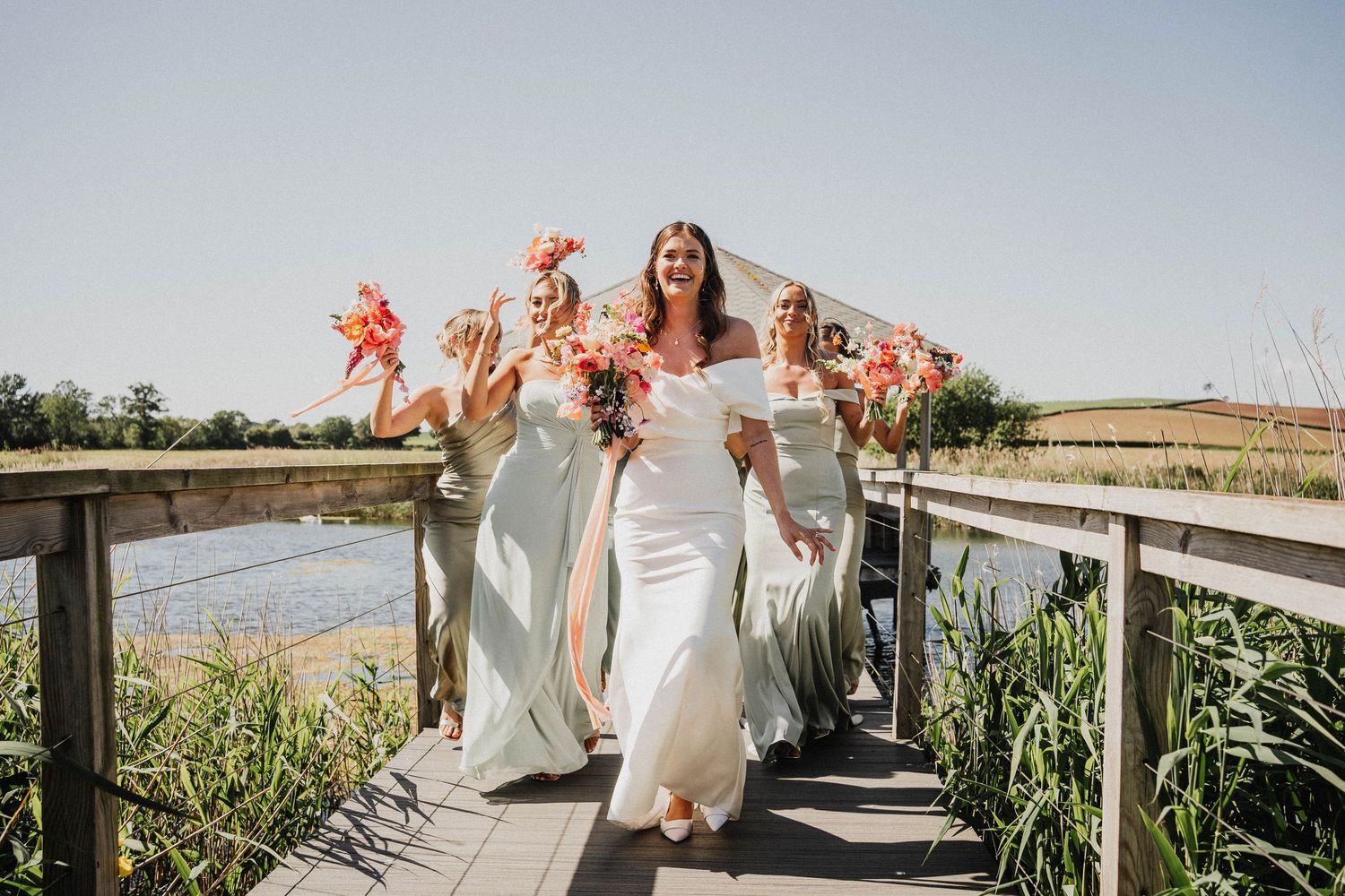 Person in a flowing white wedding dress walks along a sunlit pathway through a cornfield on a bright summer day.