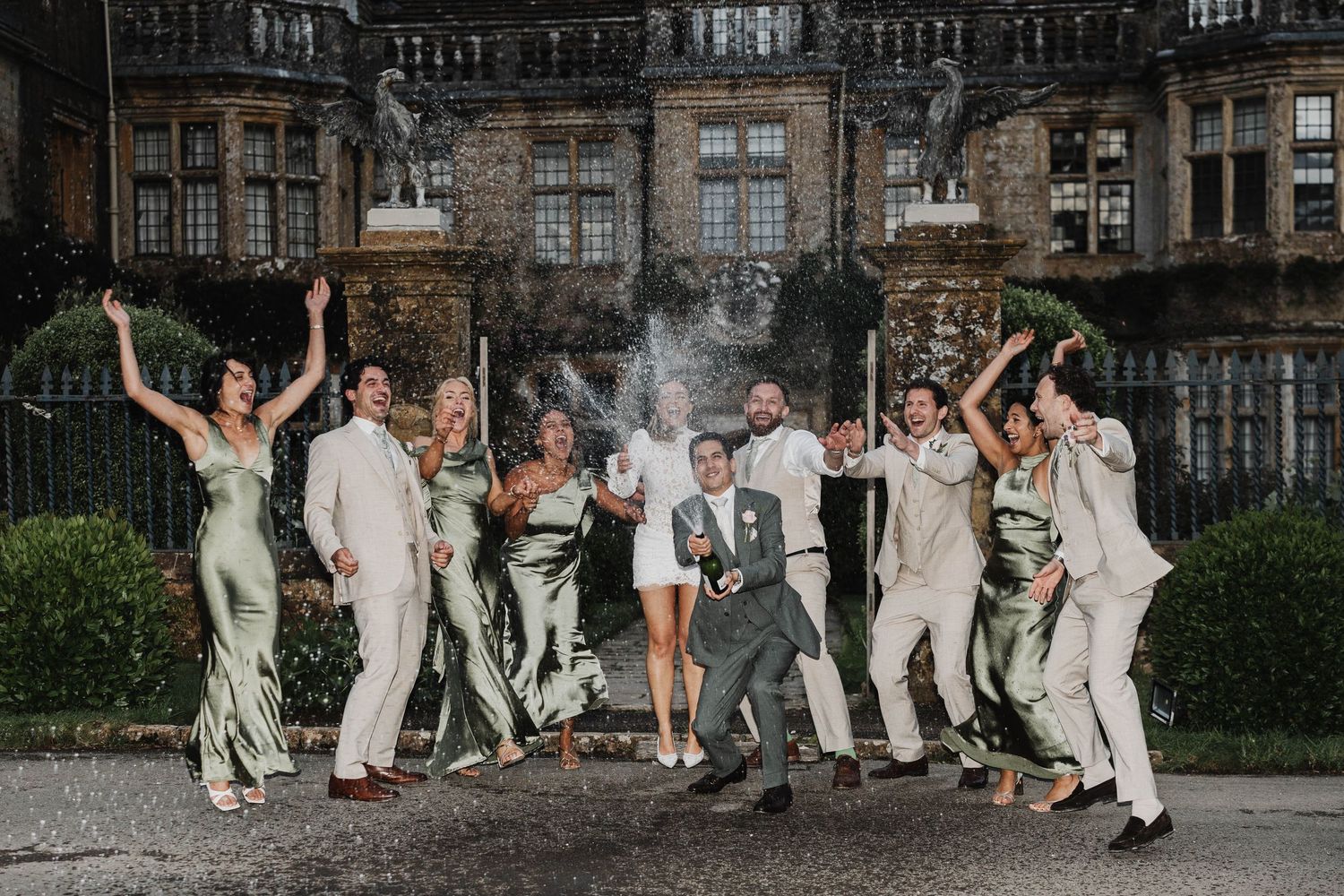 Wedding party celebrating outdoors in sage green dresses and tan suits, popping champagne in front of a grand historic venue.