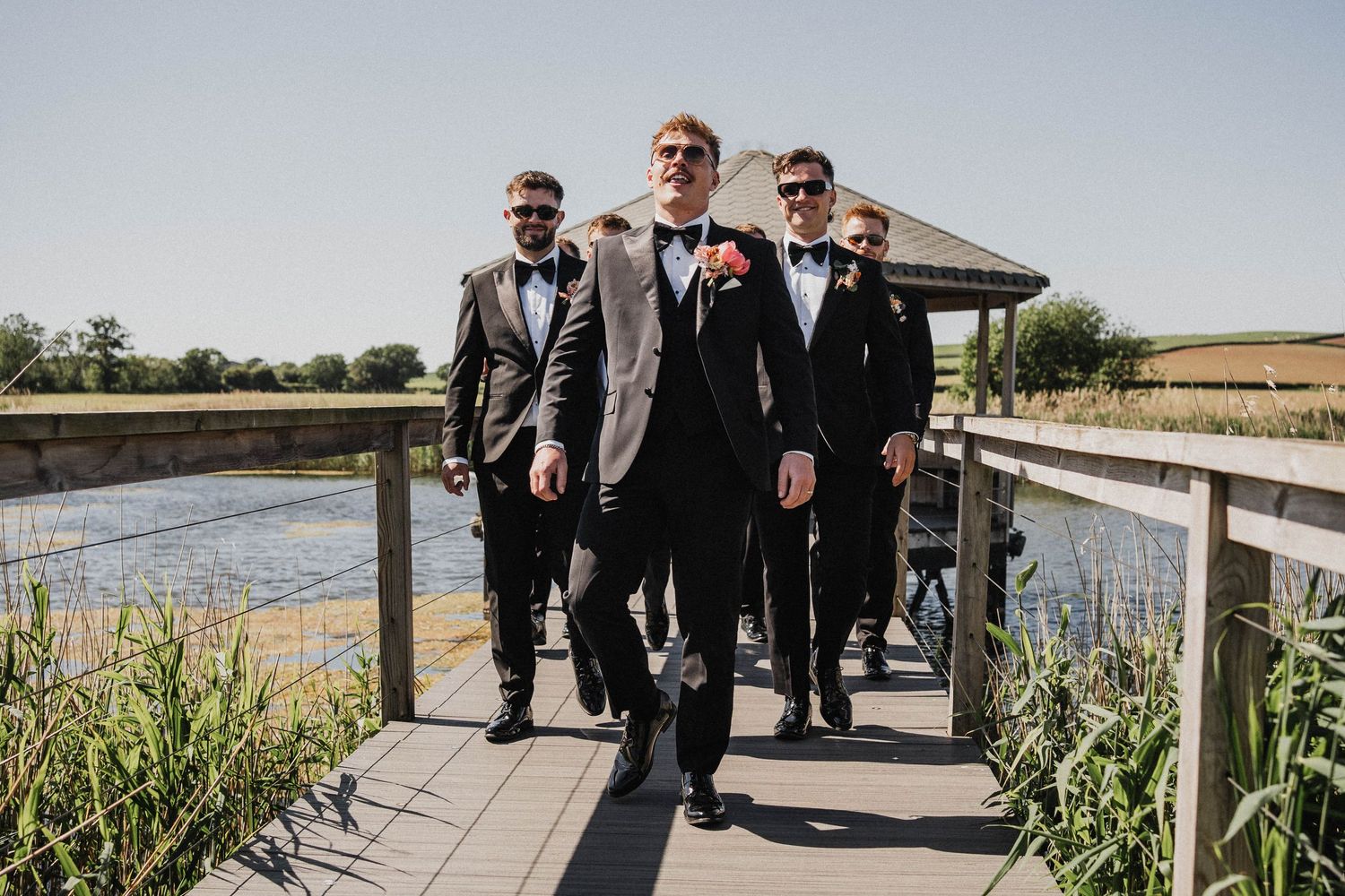 Four groomsmen in formal suits walking on a wooden boardwalk over marsh waters on a bright sunny day.