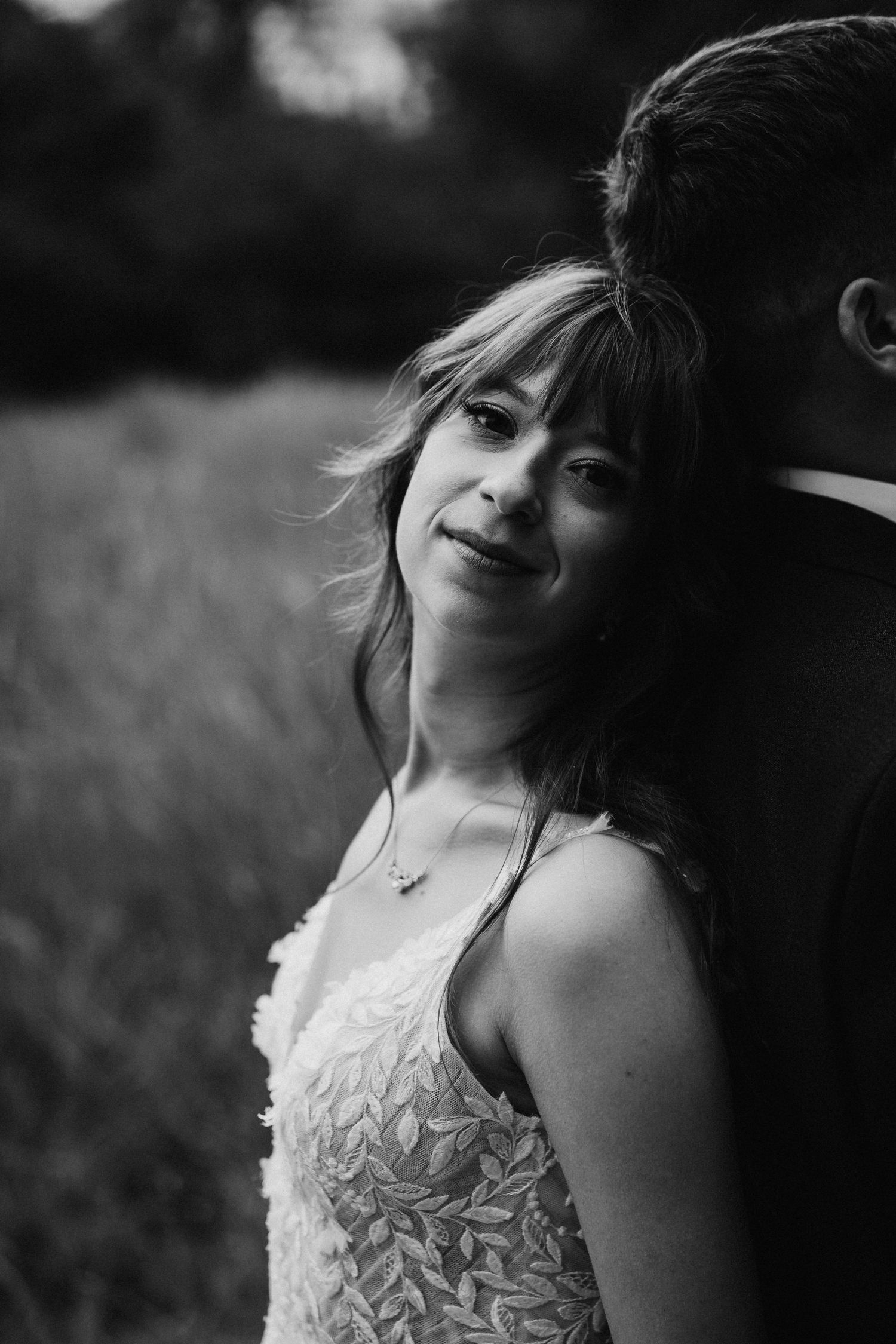 A black and white portrait of a bride in a lace dress smiling softly against a blurred outdoor background.