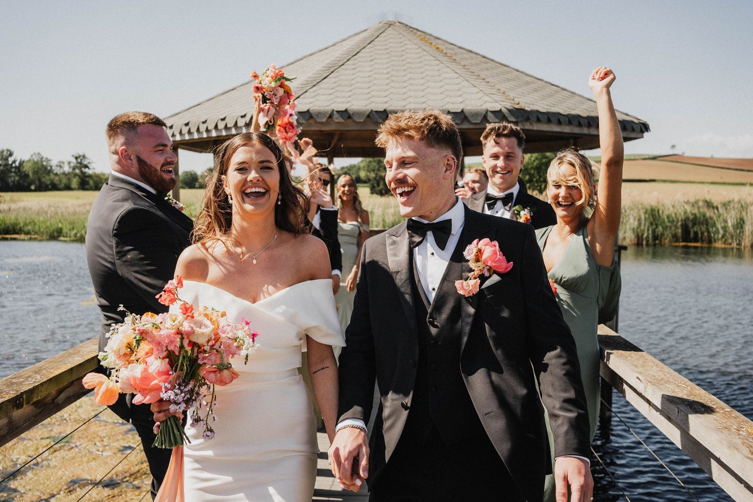 Wedding couple and bridal party celebrate on wooden dock near scenic waterfront surrounded by fall flowers and decor.