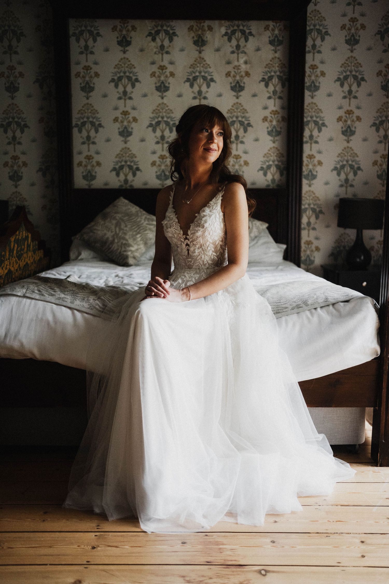 A bride in a white lace gown sits on the edge of a bed with vintage wallpaper backdrop in moody lighting.