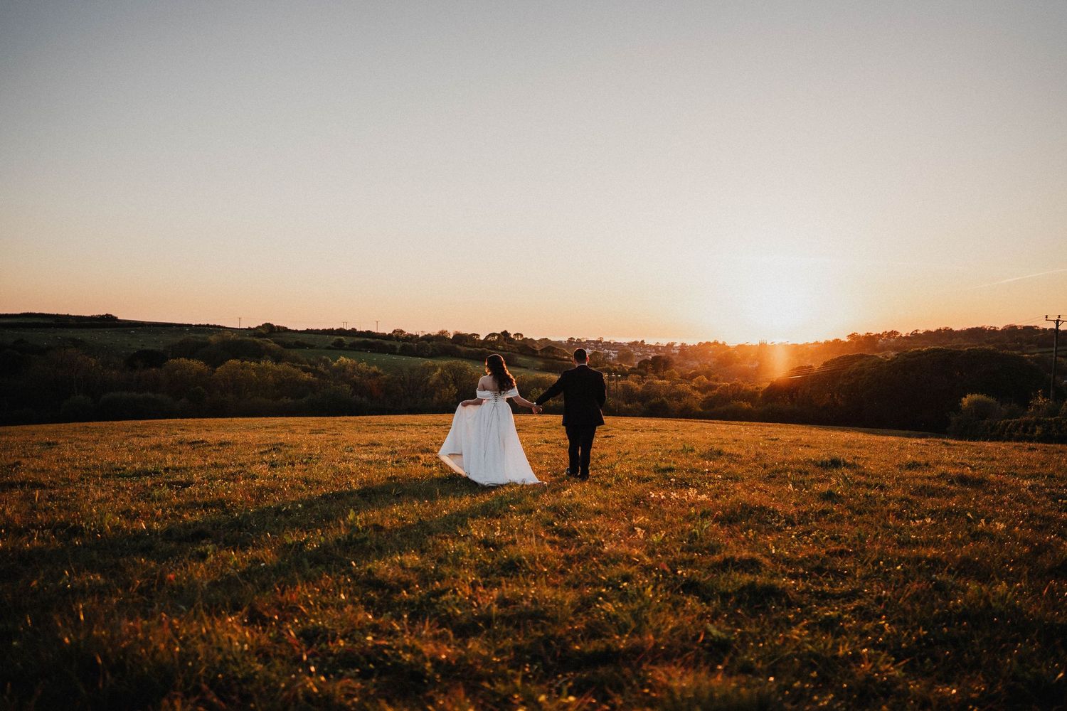 Silhouetted couple stands in grassy field during golden sunset over rolling hills.
