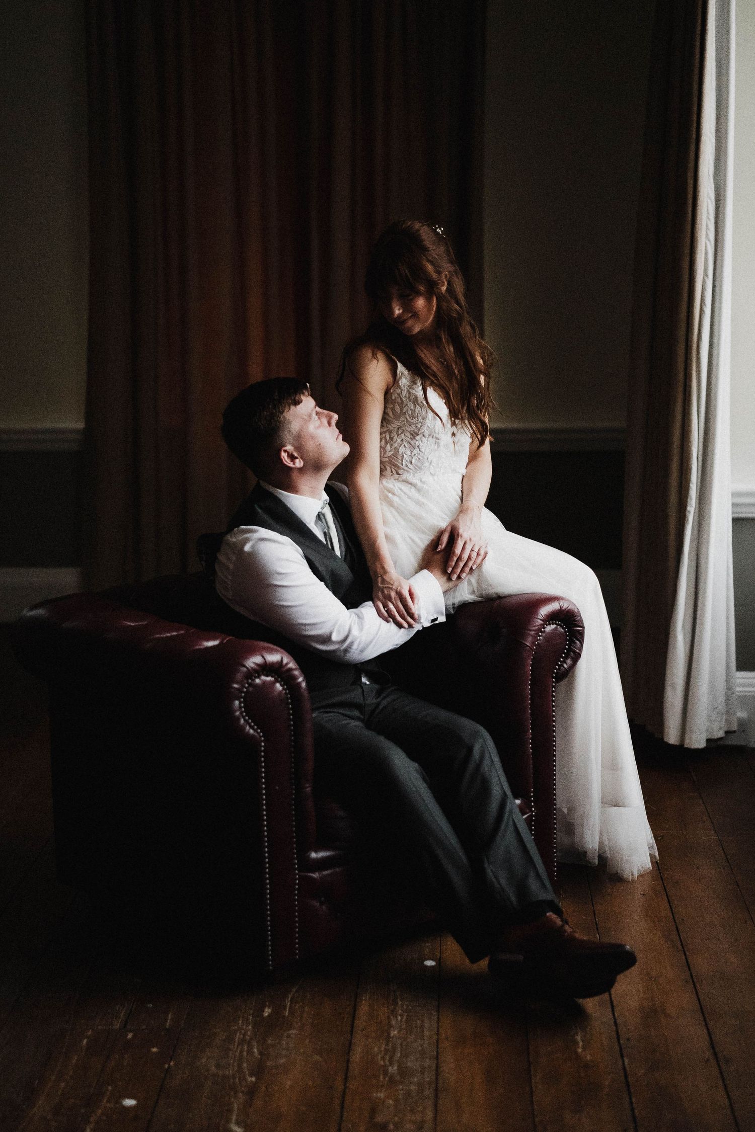 Intimate wedding portrait of couple in formal attire posing on a burgundy leather armchair.