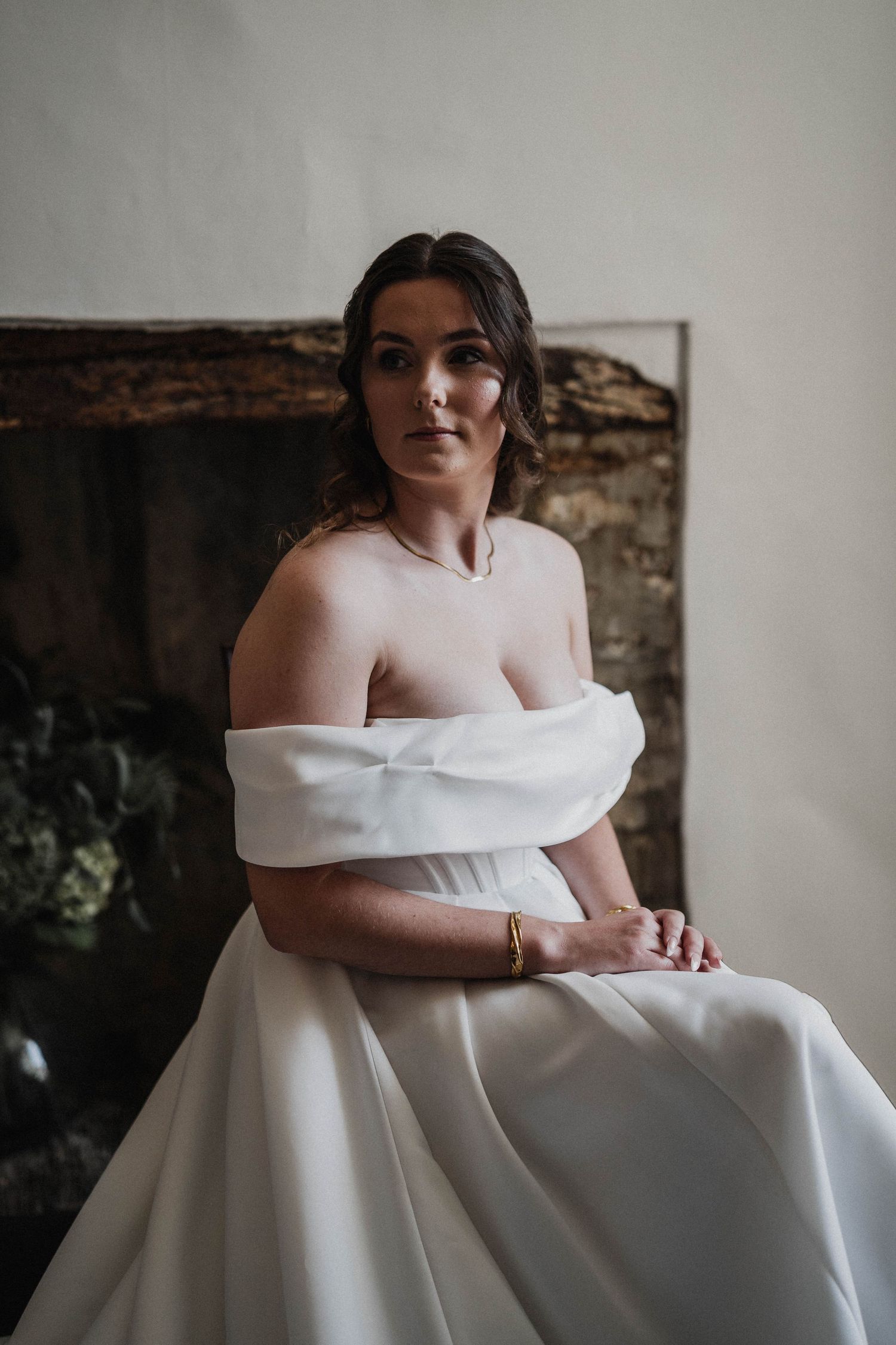 A romantic bridal portrait shows someone in an off-shoulder white wedding gown against a rustic stone wall background.