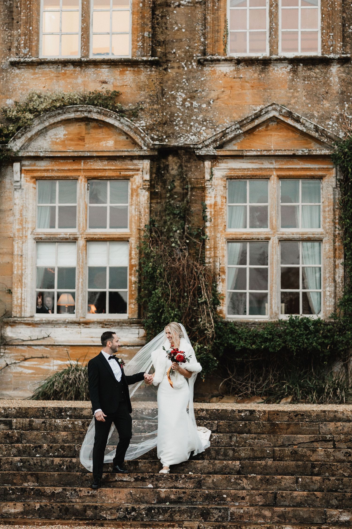 A couple poses playfully on stone steps against a historic building facade with large windows.