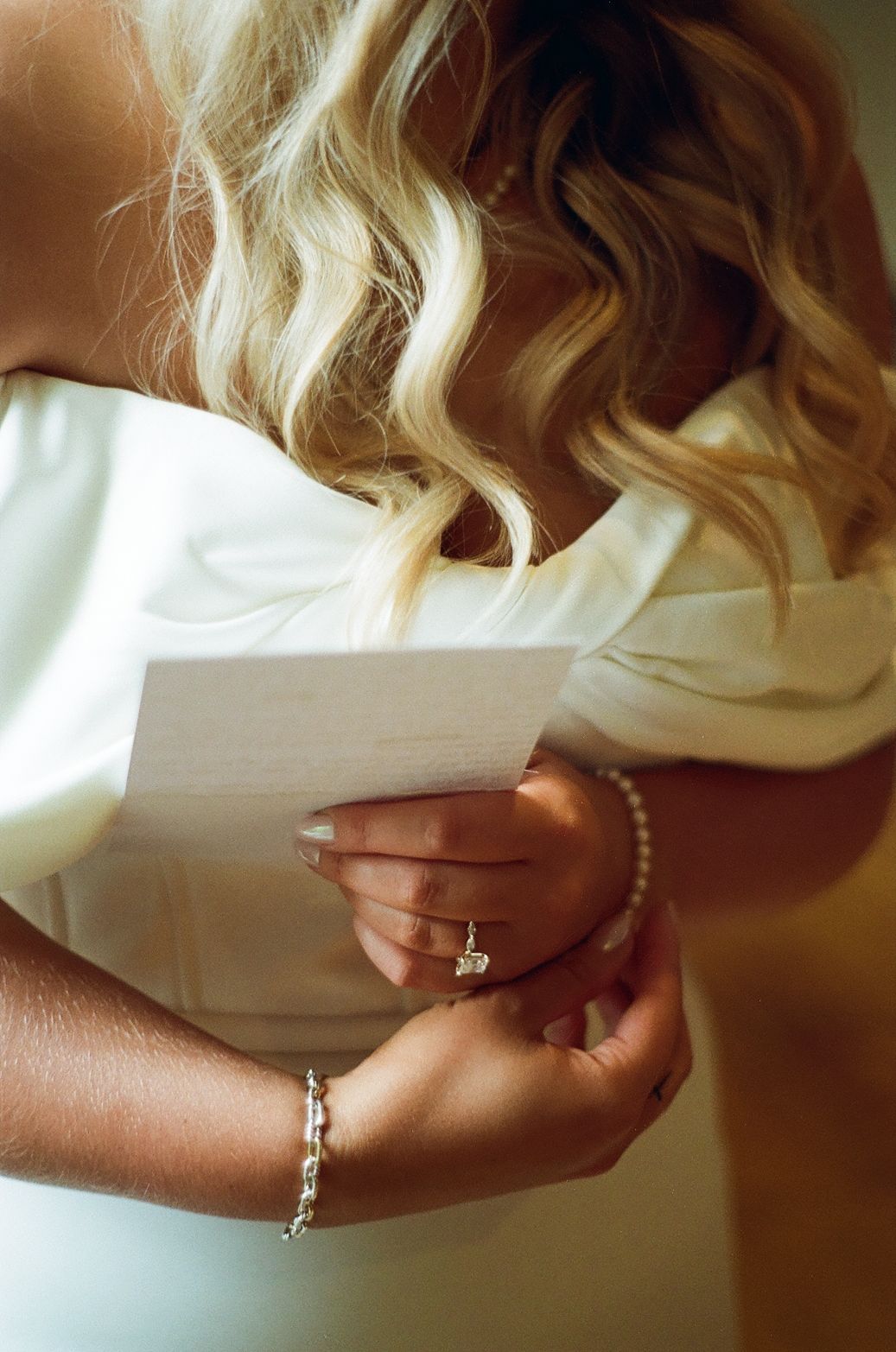 Close-up of hands holding a letter, with blonde wavy hair visible and wearing a pearl bracelet and engagement ring.