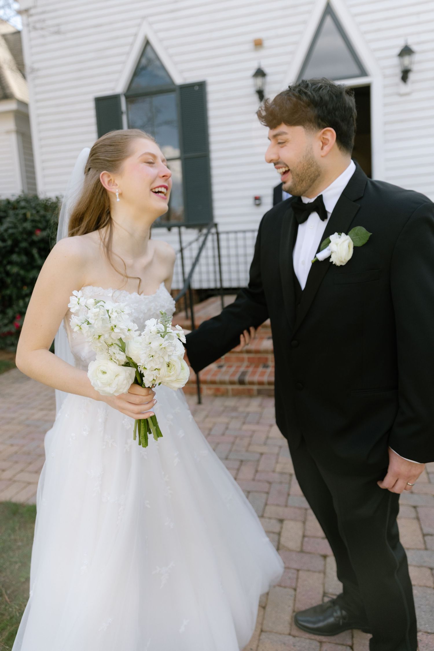 A bride and groom share joyful laughter together during their wedding photo session on a brick patio.