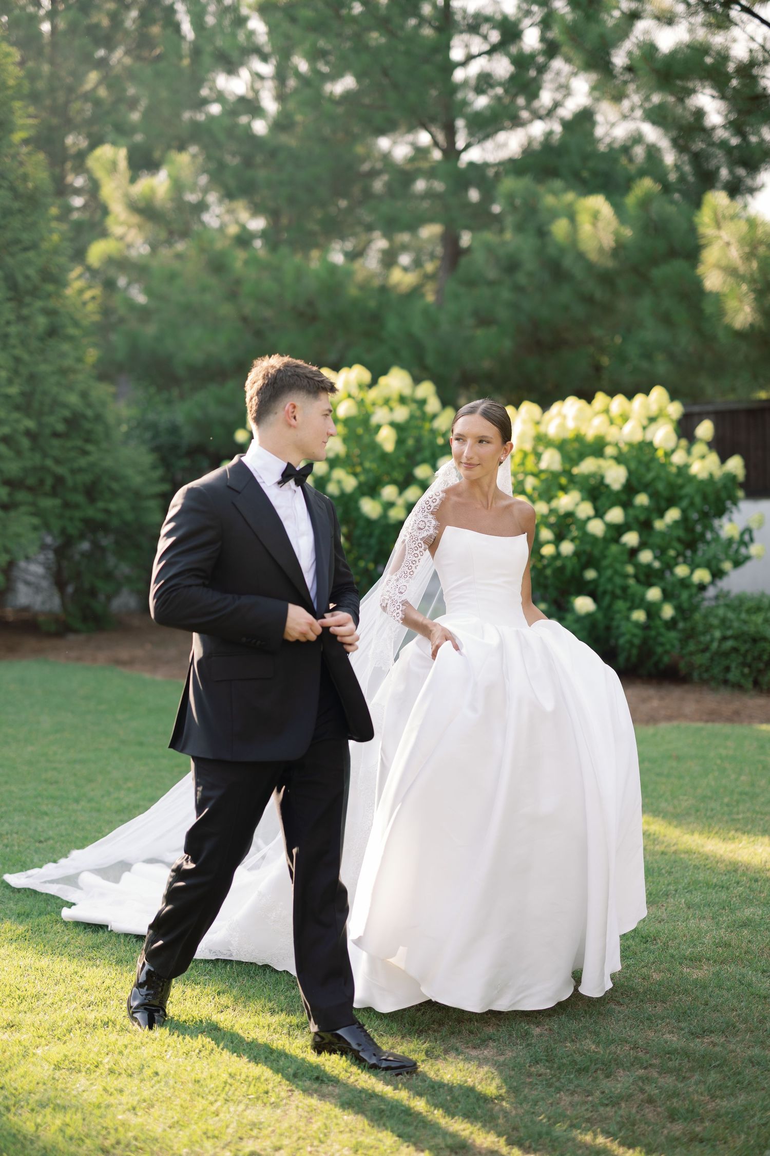 Bride in strapless ball gown and groom in black tuxedo share a romantic moment on lush green lawn.