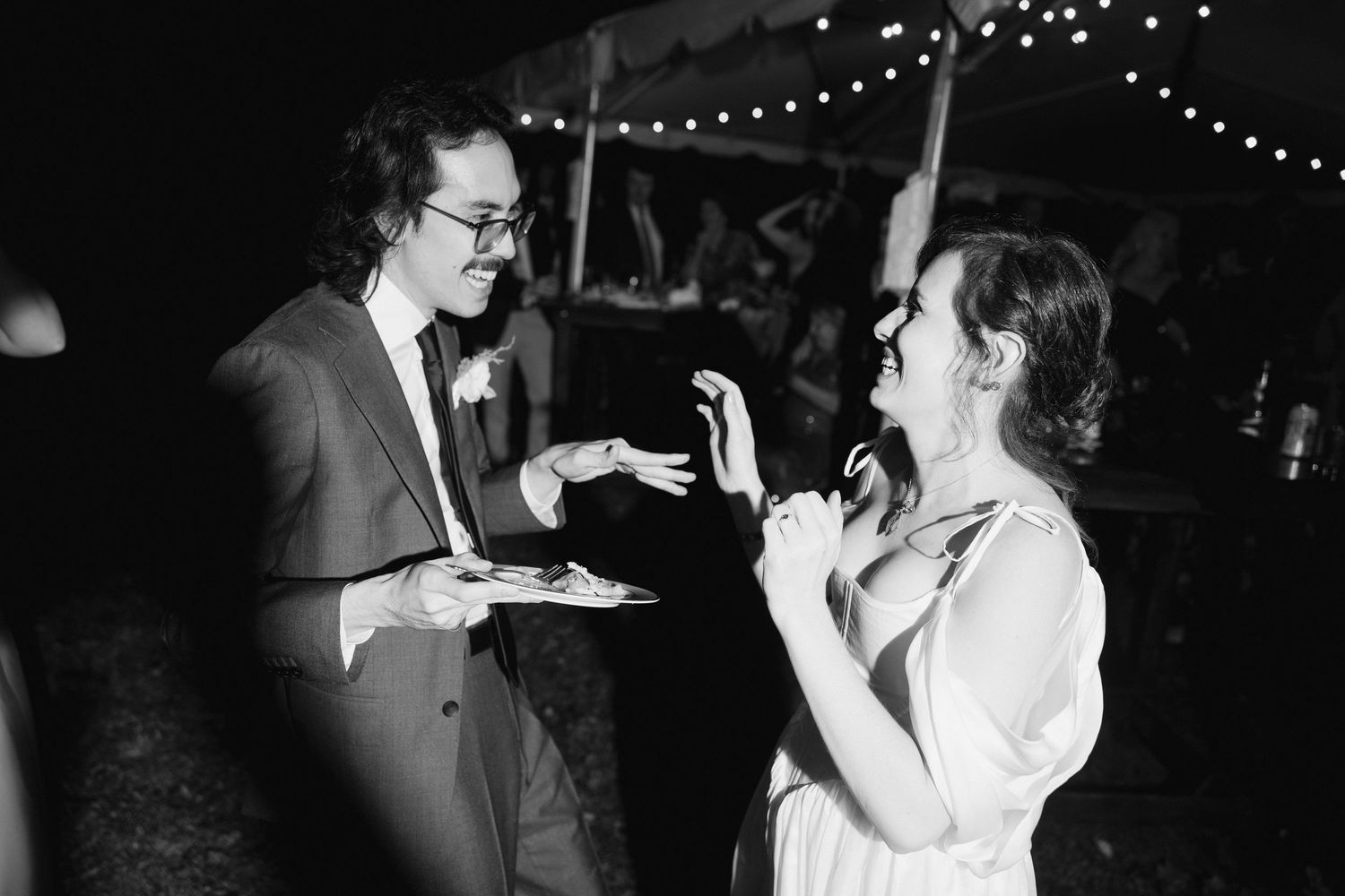 A black and white photo of a couple sharing cake at a wedding reception with string lights in the background.