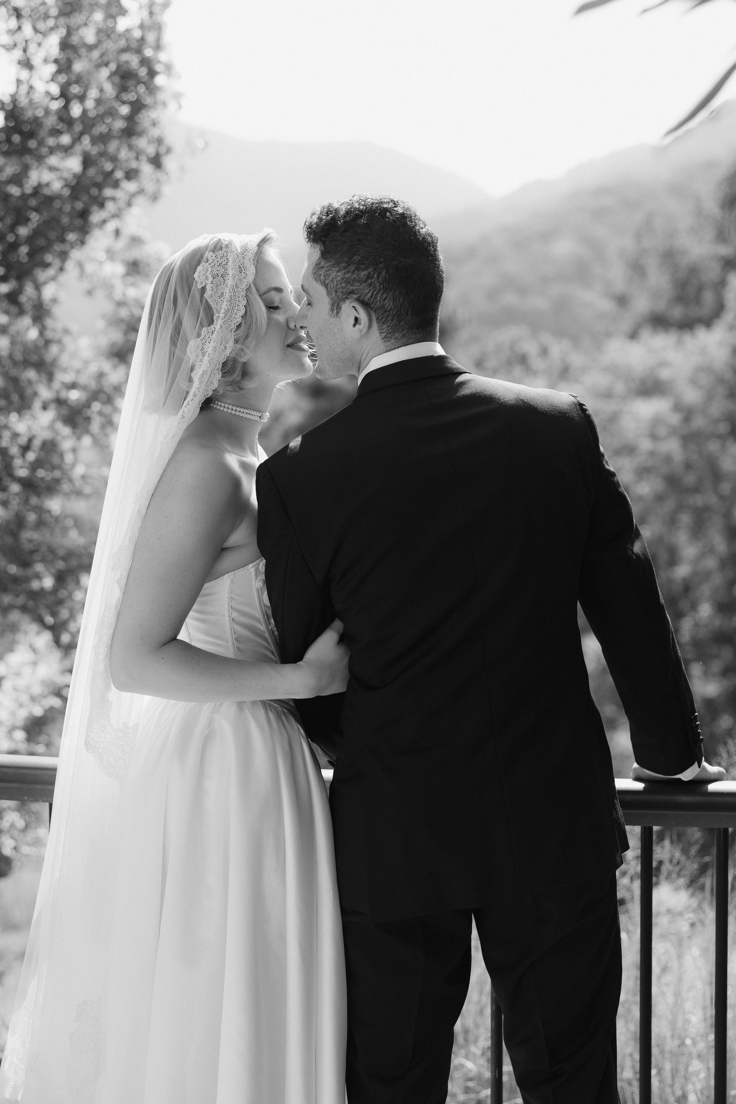 Black and white photo of a bride and groom sharing a romantic moment on a balcony with mountain views.