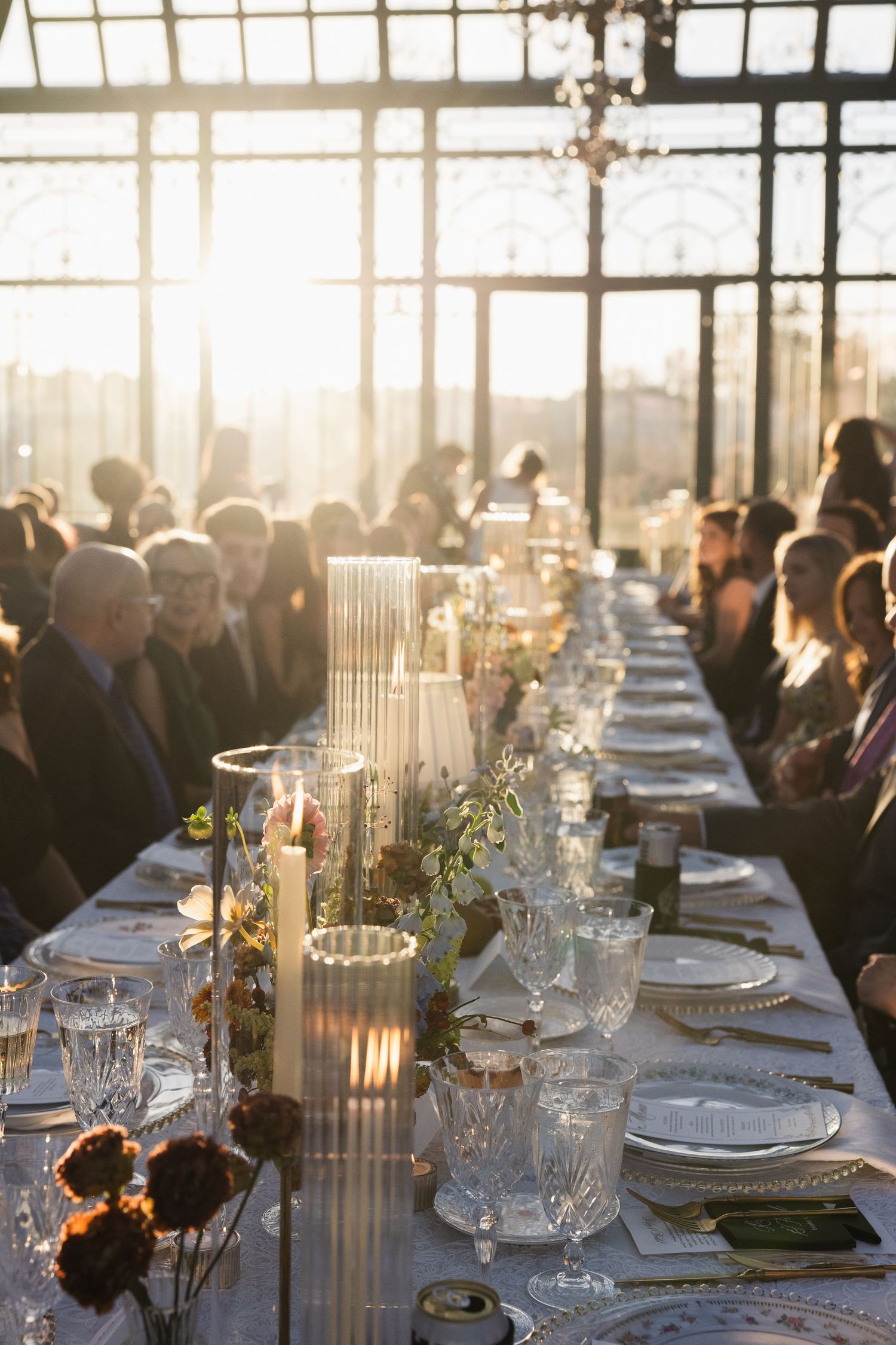 Elegant wedding reception table setting with glowing candles and floral centerpieces at sunset in a glass-enclosed venue.