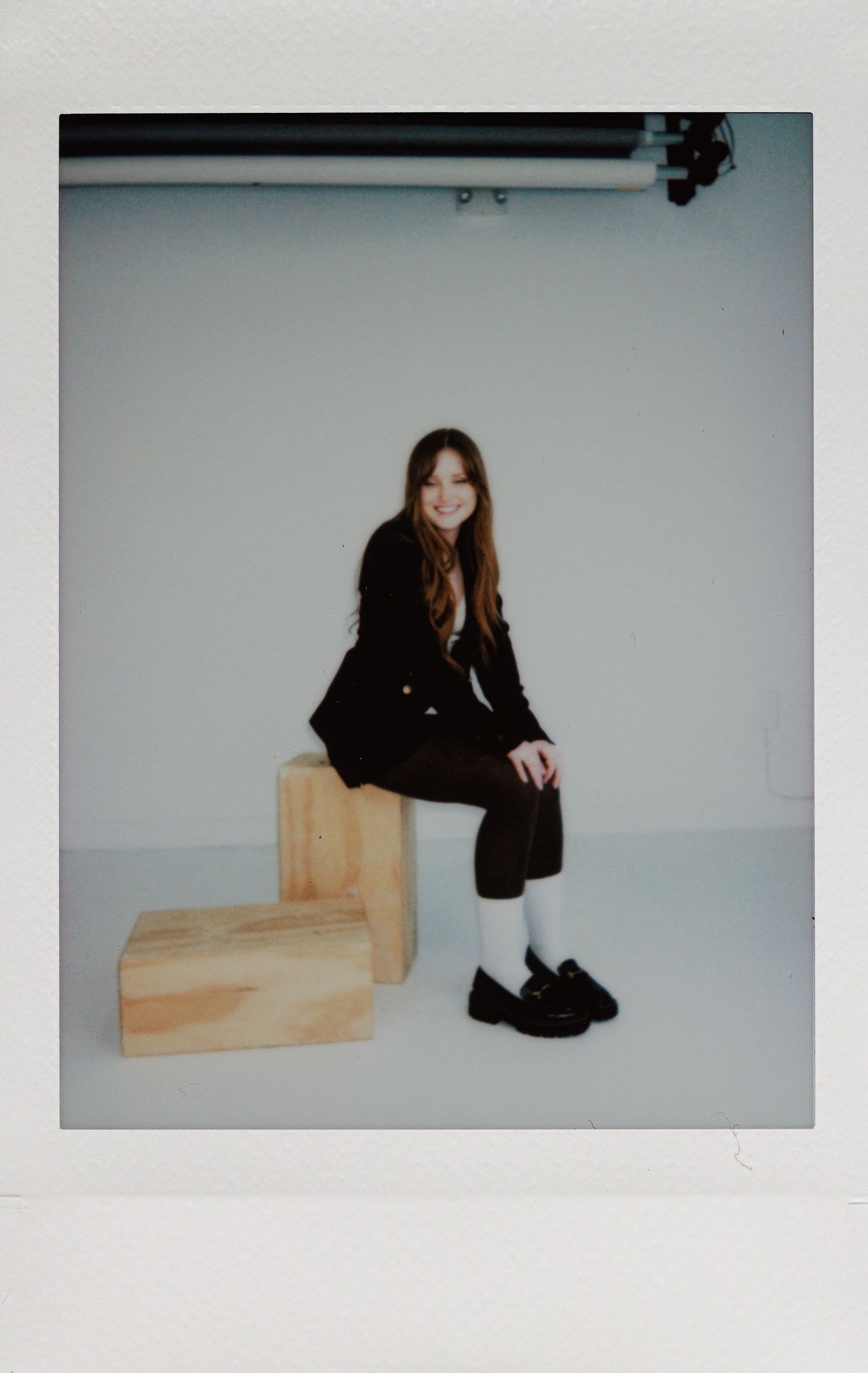 A person in a black outfit sits on wooden blocks against a white studio backdrop wearing chunky black shoes.
