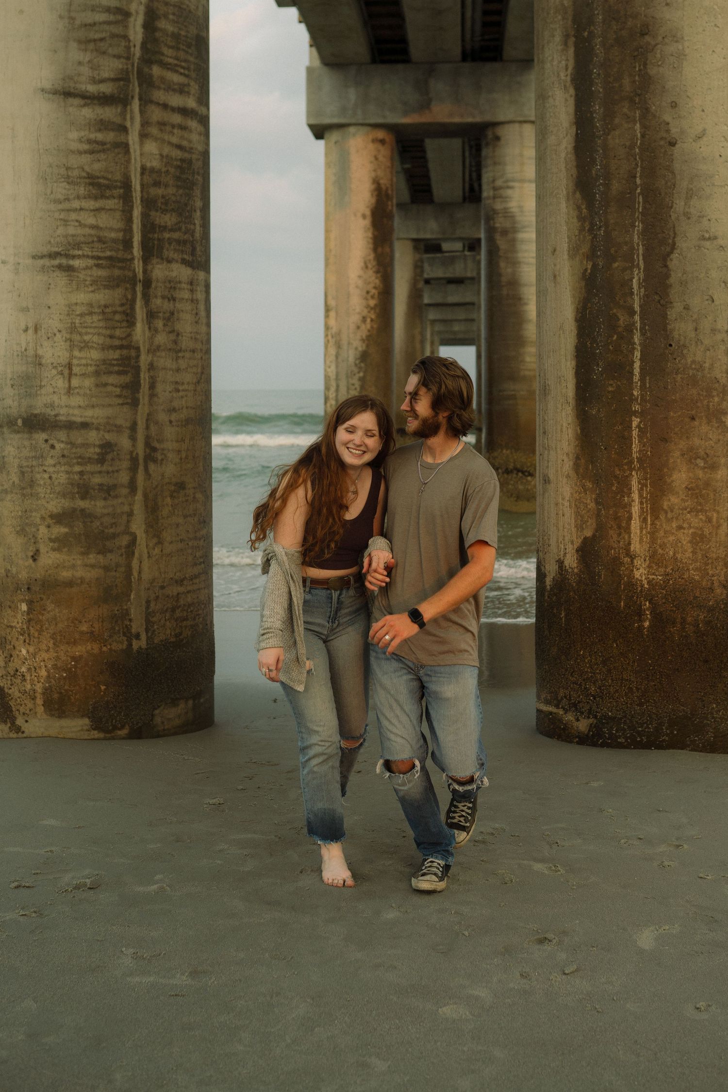 Couple standing between pier columns at beach, sharing intimate moment.