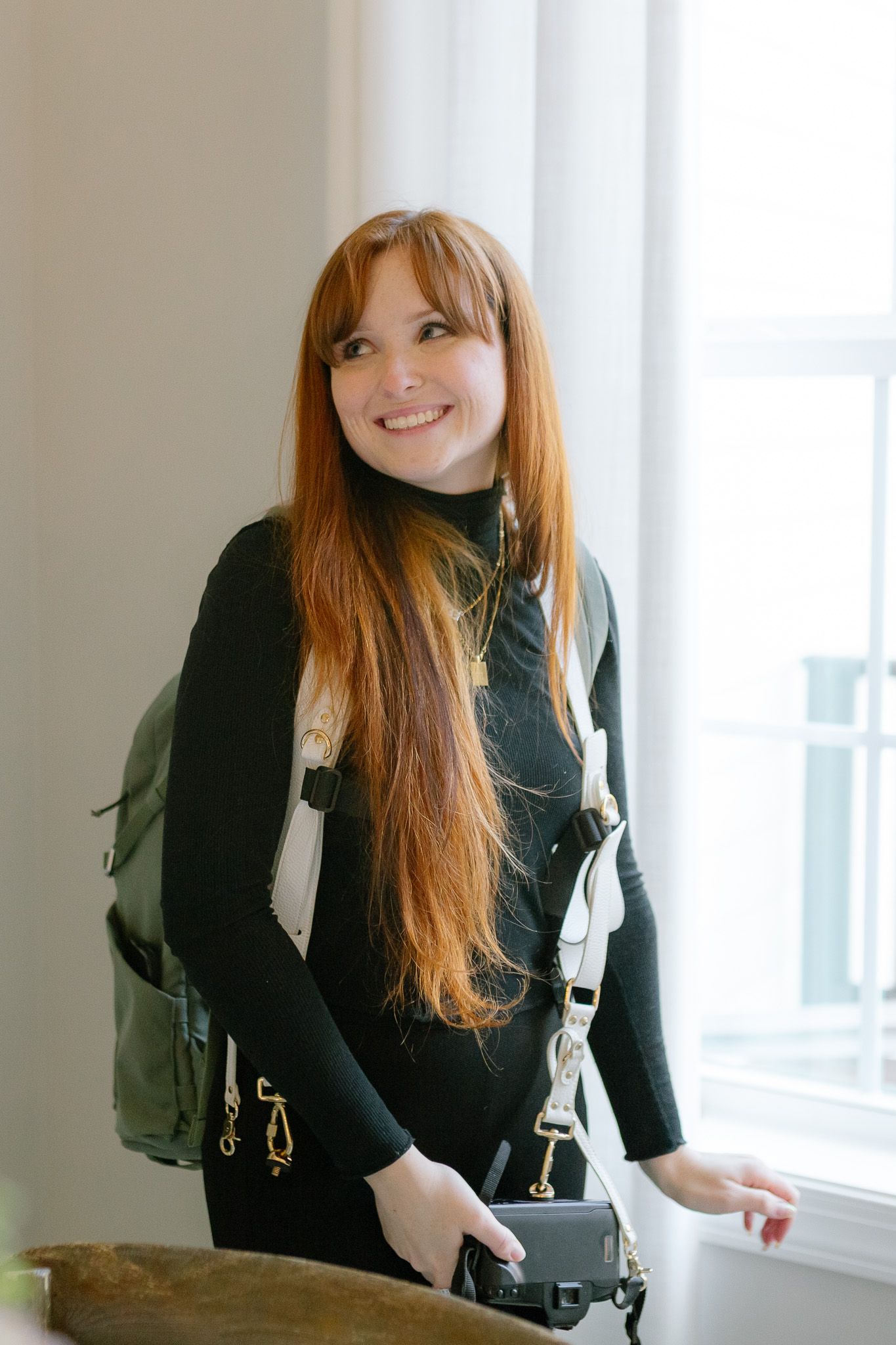 A person with long red hair wearing a black sweater stands by a window in natural light.