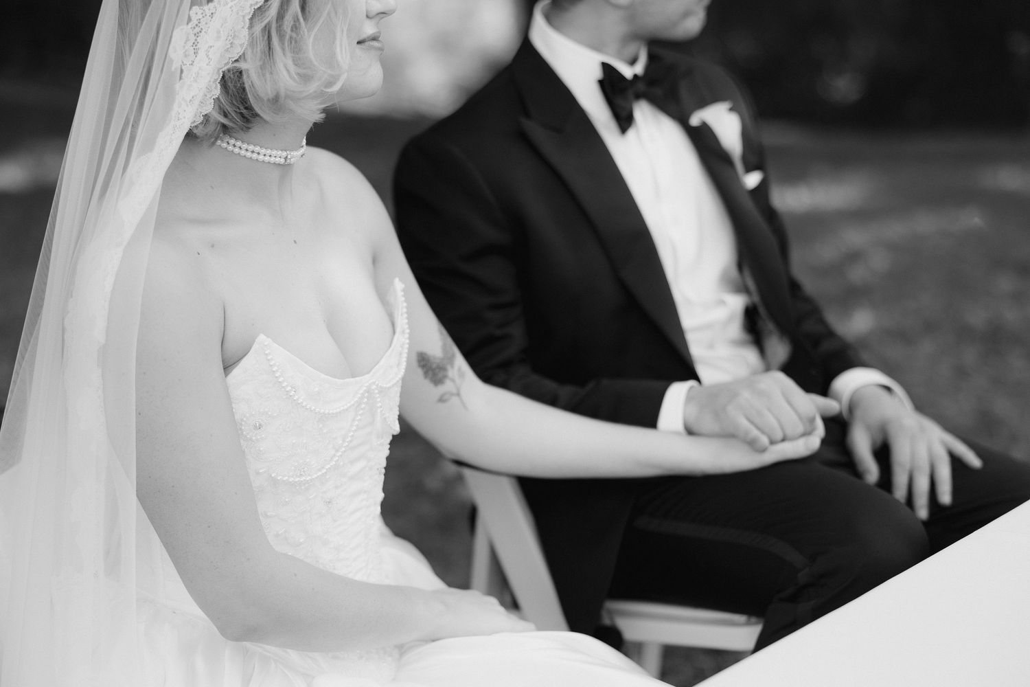 Black and white photo of bride and groom holding hands during wedding ceremony, bride wearing strapless gown and veil.