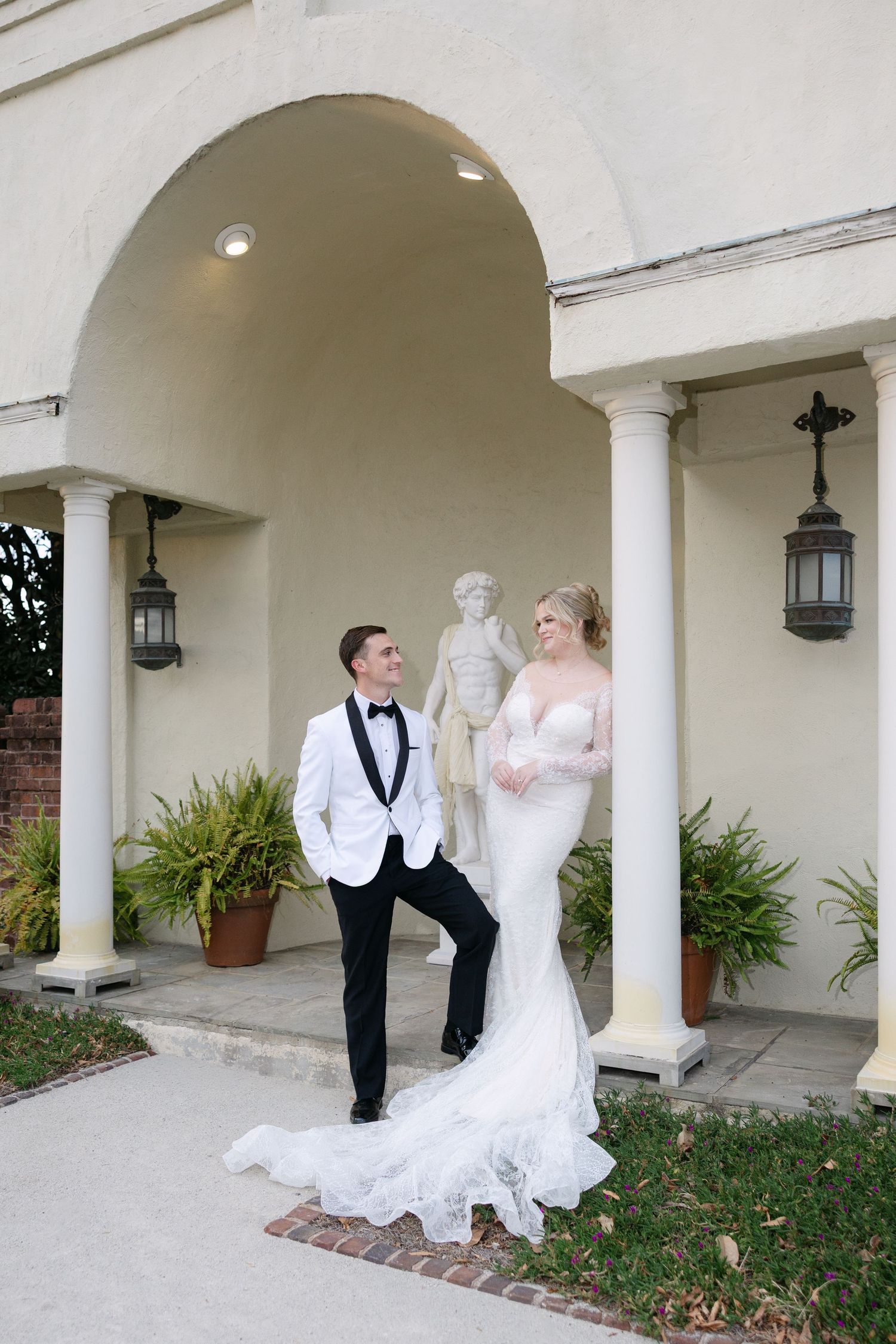 A wedding couple poses under a series of white archways with brick and stone columns in an elegant outdoor setting.