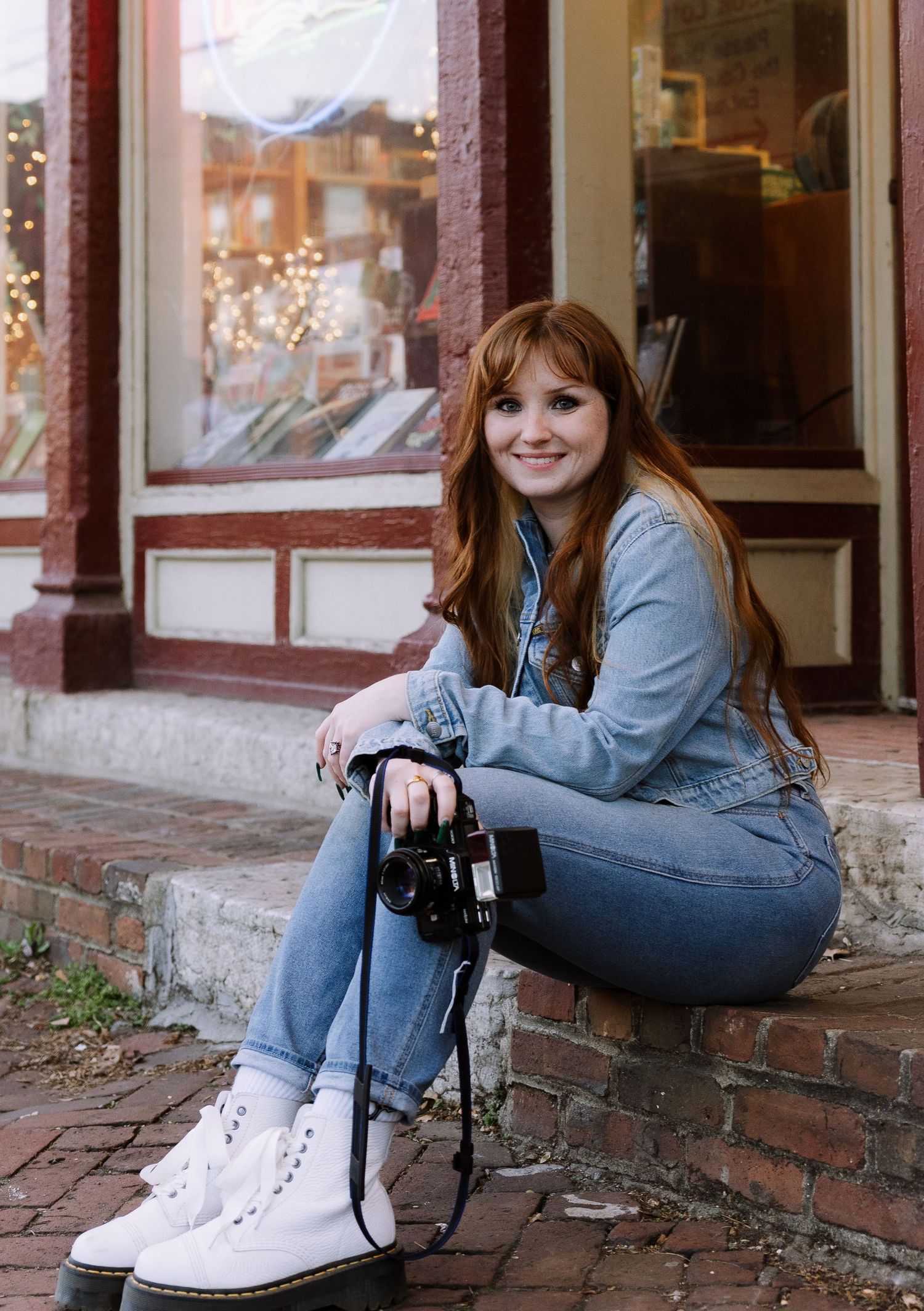 Photographer with camera sitting on brick steps outside storefront with holiday lights.