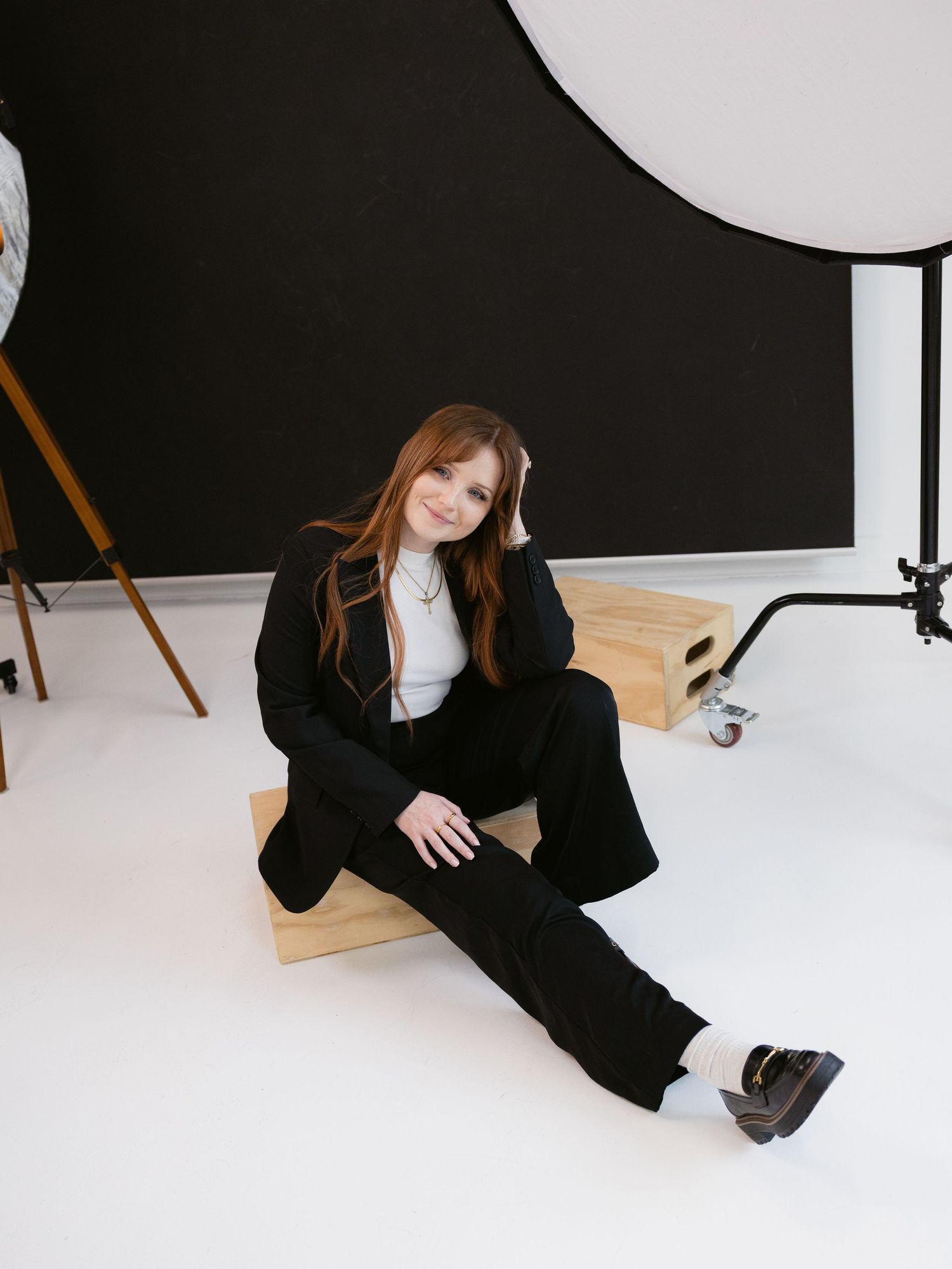 A person in a black blazer sits on a white floor in a studio setting, creating a minimalist composition.