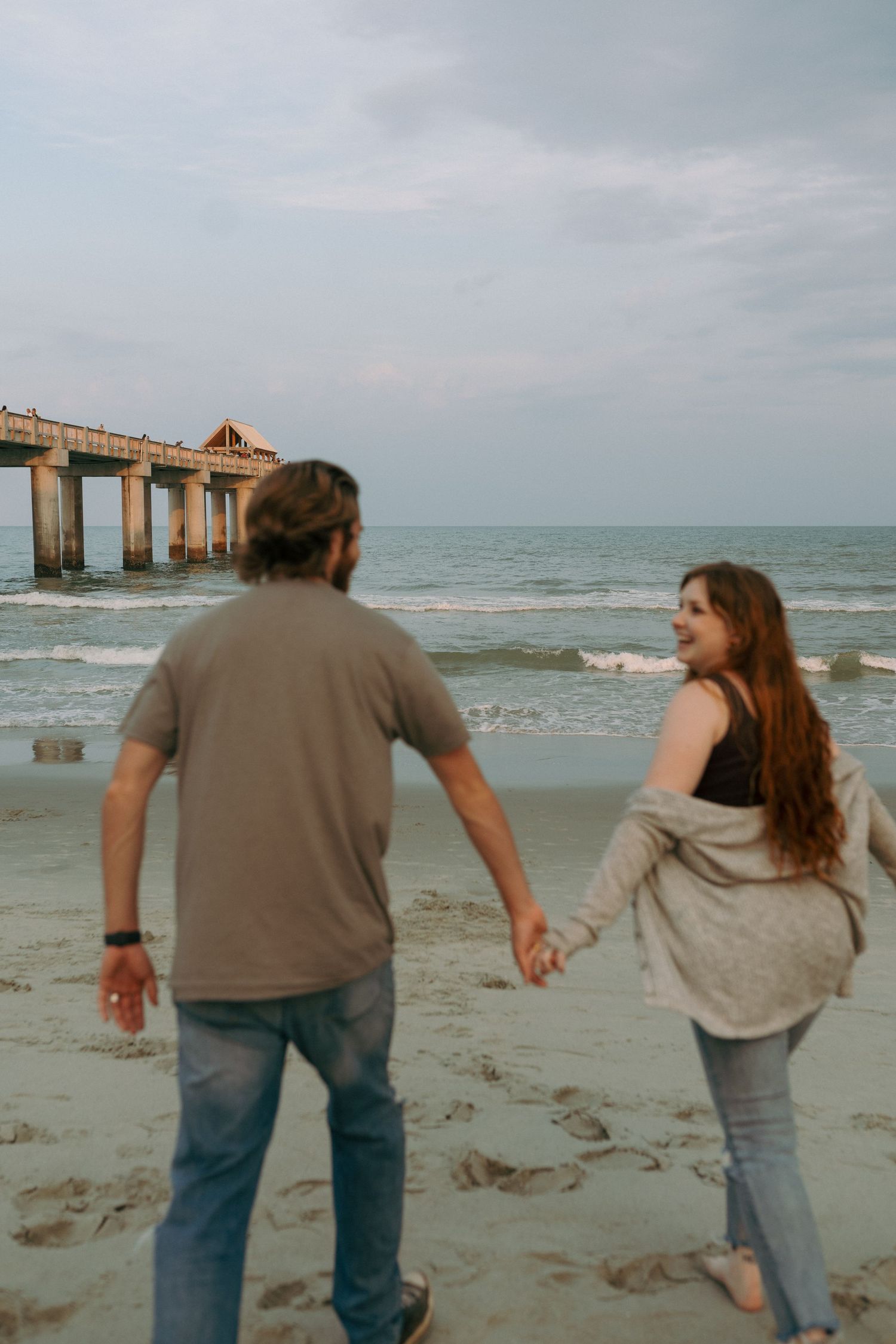 Couple holding hands while walking on beach near pier at sunset.