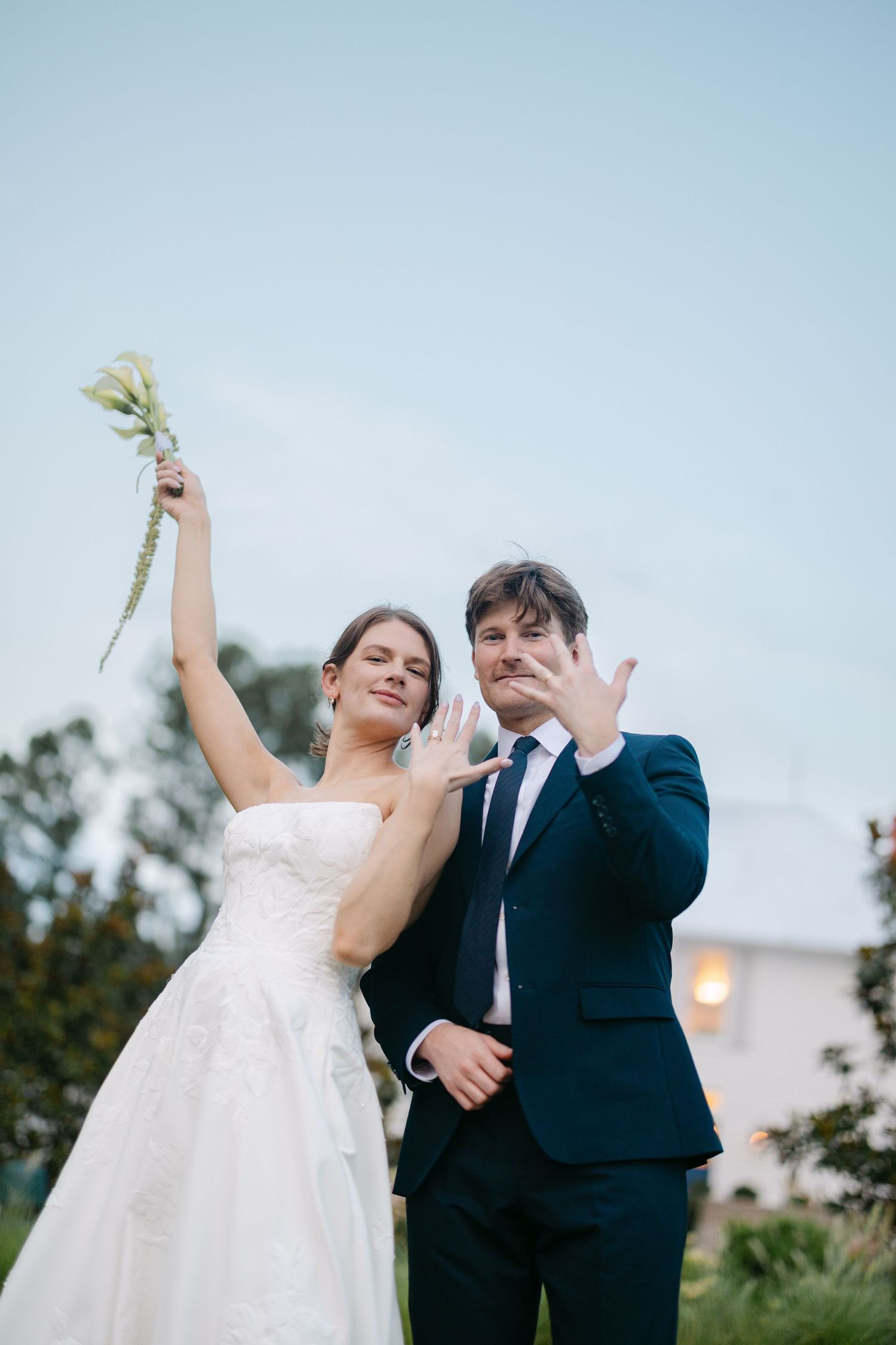 Newlyweds showing wedding rings, bride in white gown holding bouquet aloft, groom in navy suit, outdoors at dusk.