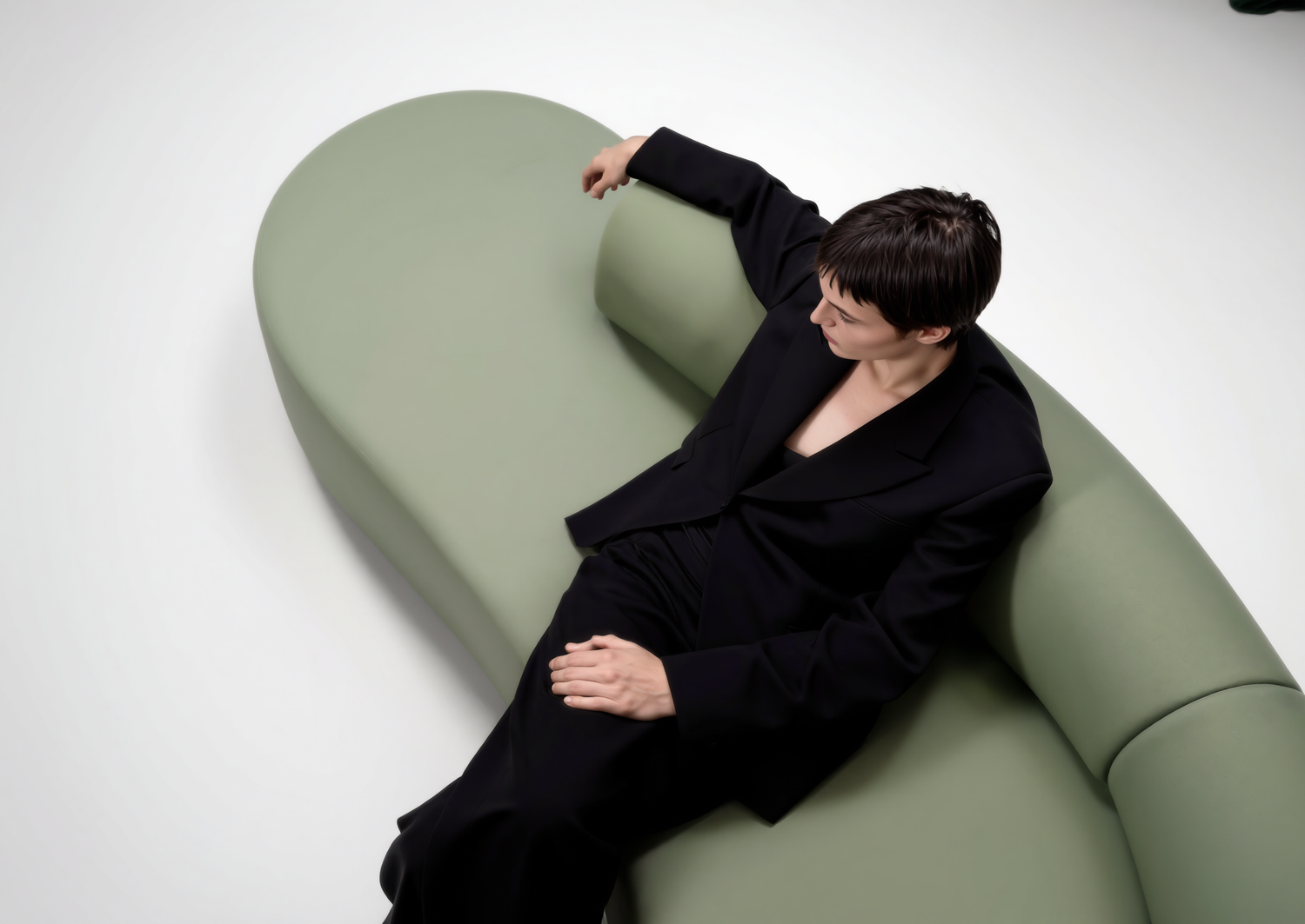 Woman in black outfit reclines on a curved sage green modern sofa against a white background.