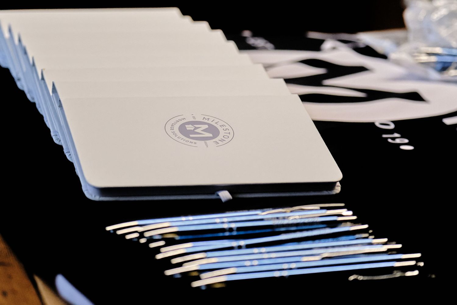Stack of white notebooks with embossed logo beside branded pens on a dark table at a corporate event.